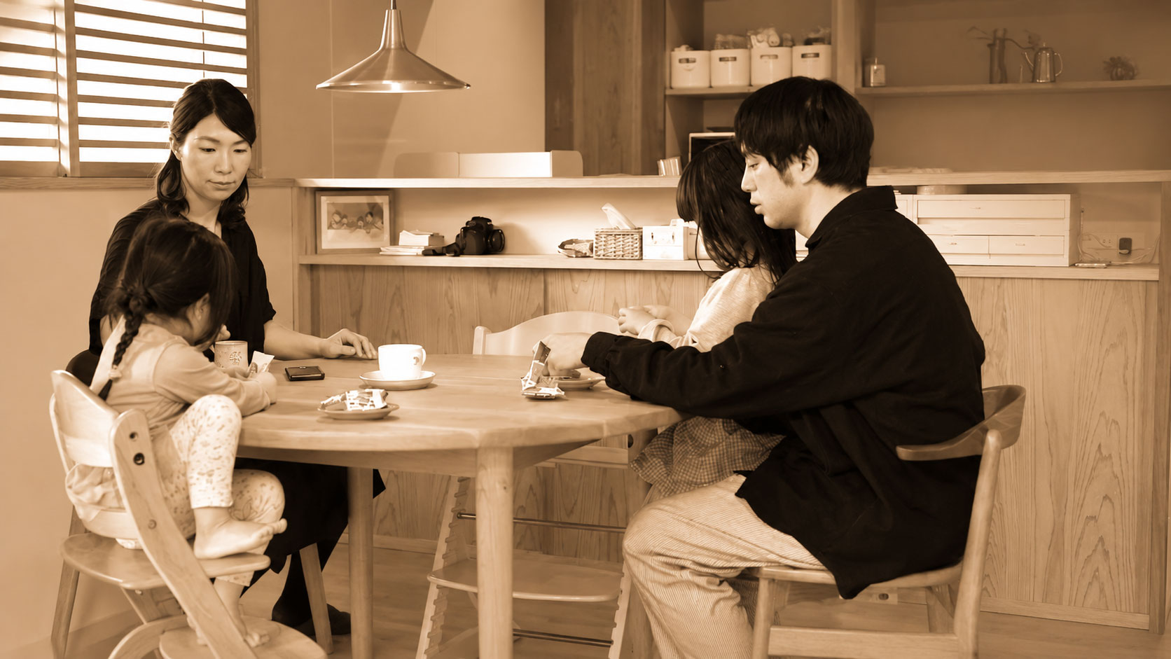 Family sits around a wooden table in a warm kitchen, sharing tea and snacks together.
