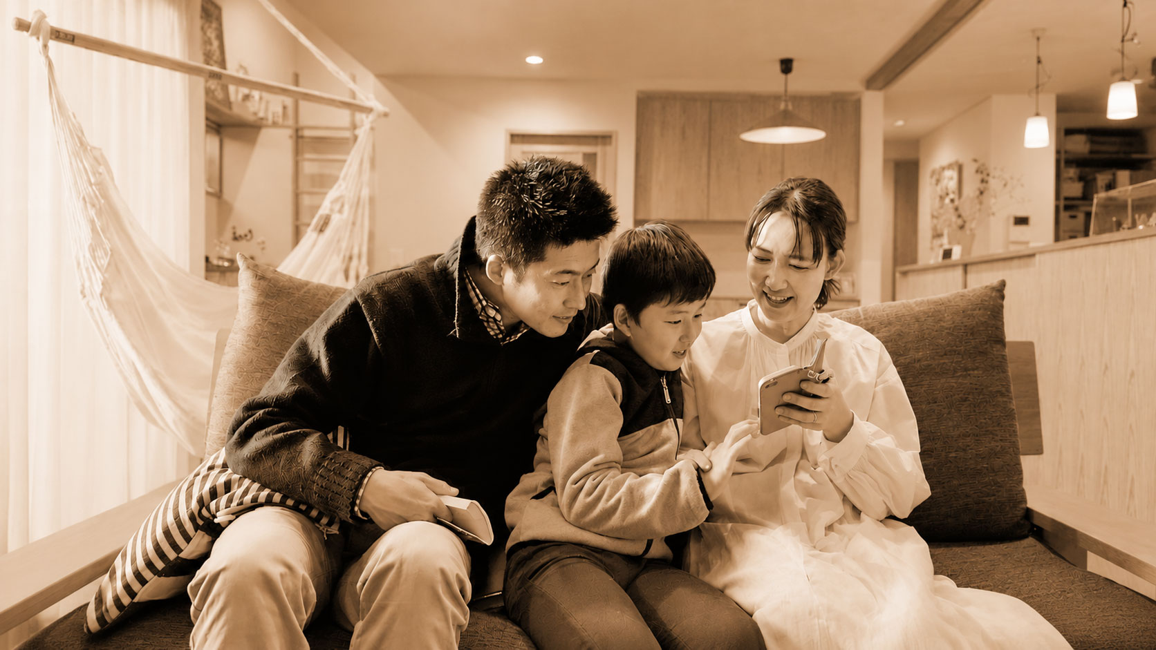 Family of three (father, child, mother) smiles as they look at a smartphone together on a sofa in a cozy living space.