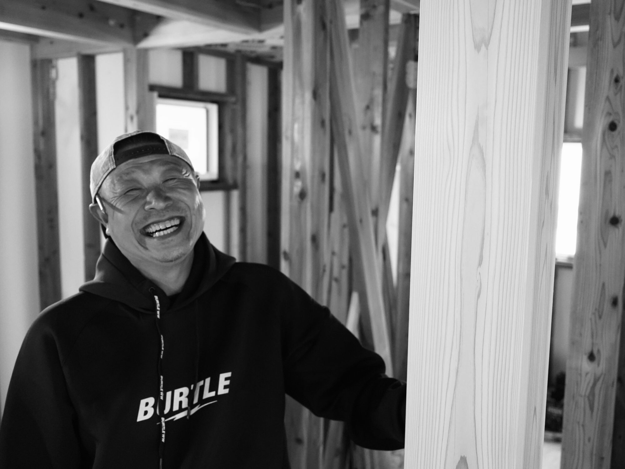 Smiling man in a backward cap and Bur tle hoodie stands among wooden framing in a new construction site.