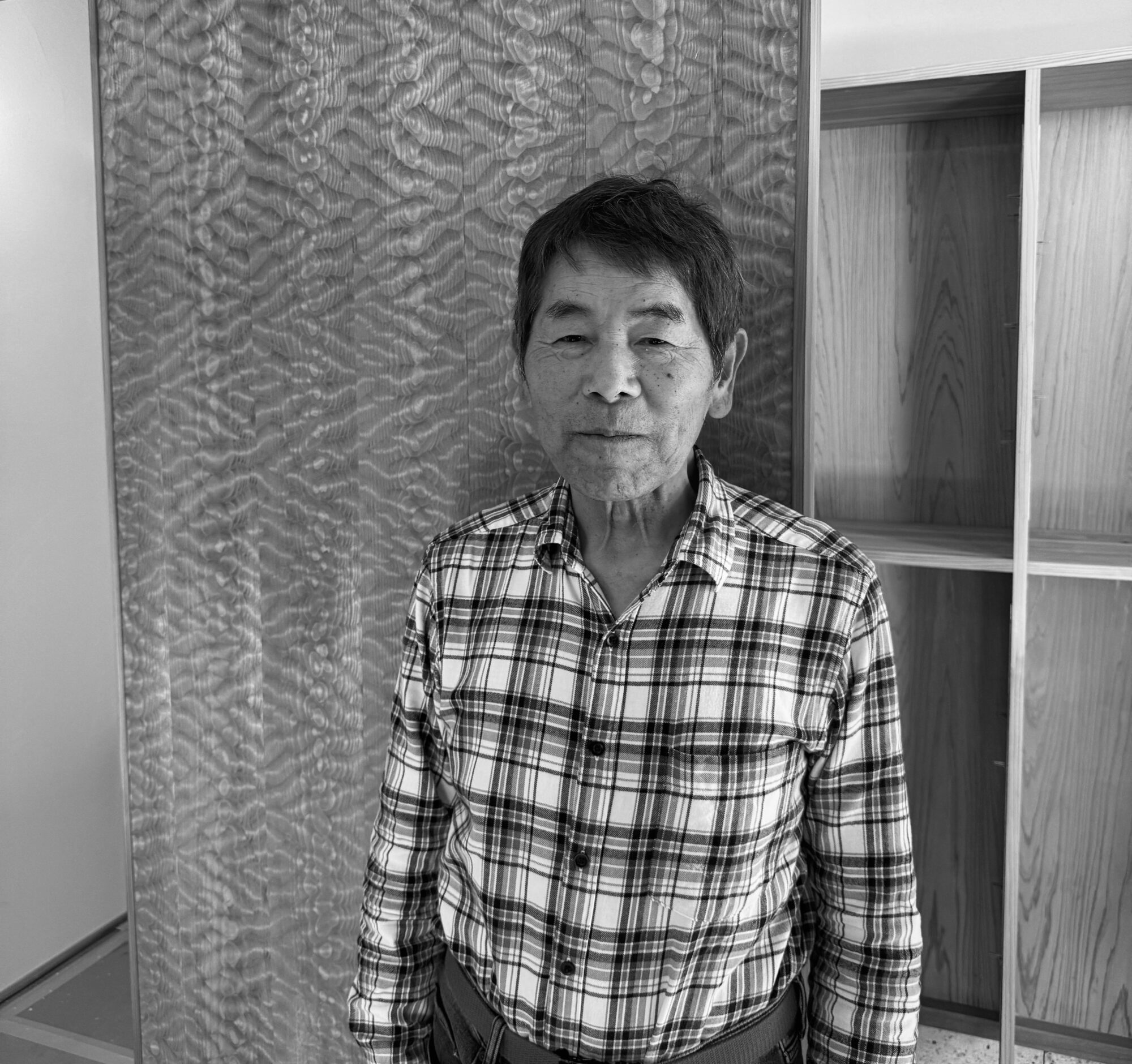 Older man in a plaid shirt standing indoors in front of a textured divider and wood shelves behind him.