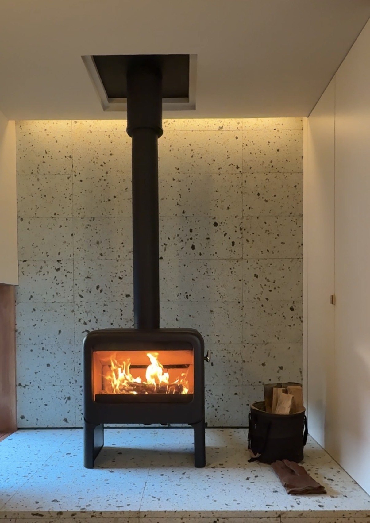 Black wood stove with glass door showing flames, pipe extending to ceiling in a minimalist room with a speckled wall behind it.