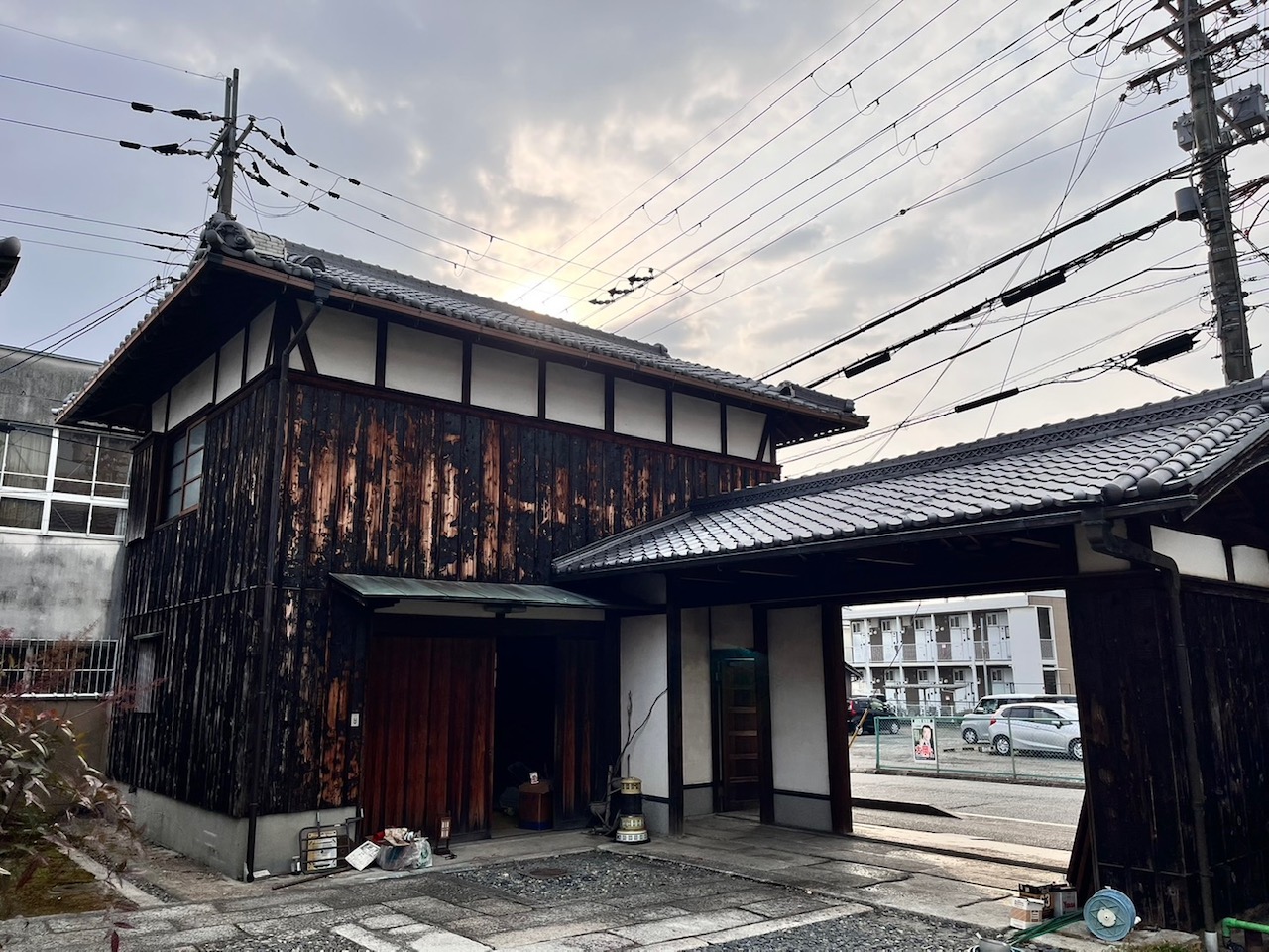 Traditional Japanese wooden building with dark weathered planks and tiled roof beside a gate, power lines overhead in an urban setting.