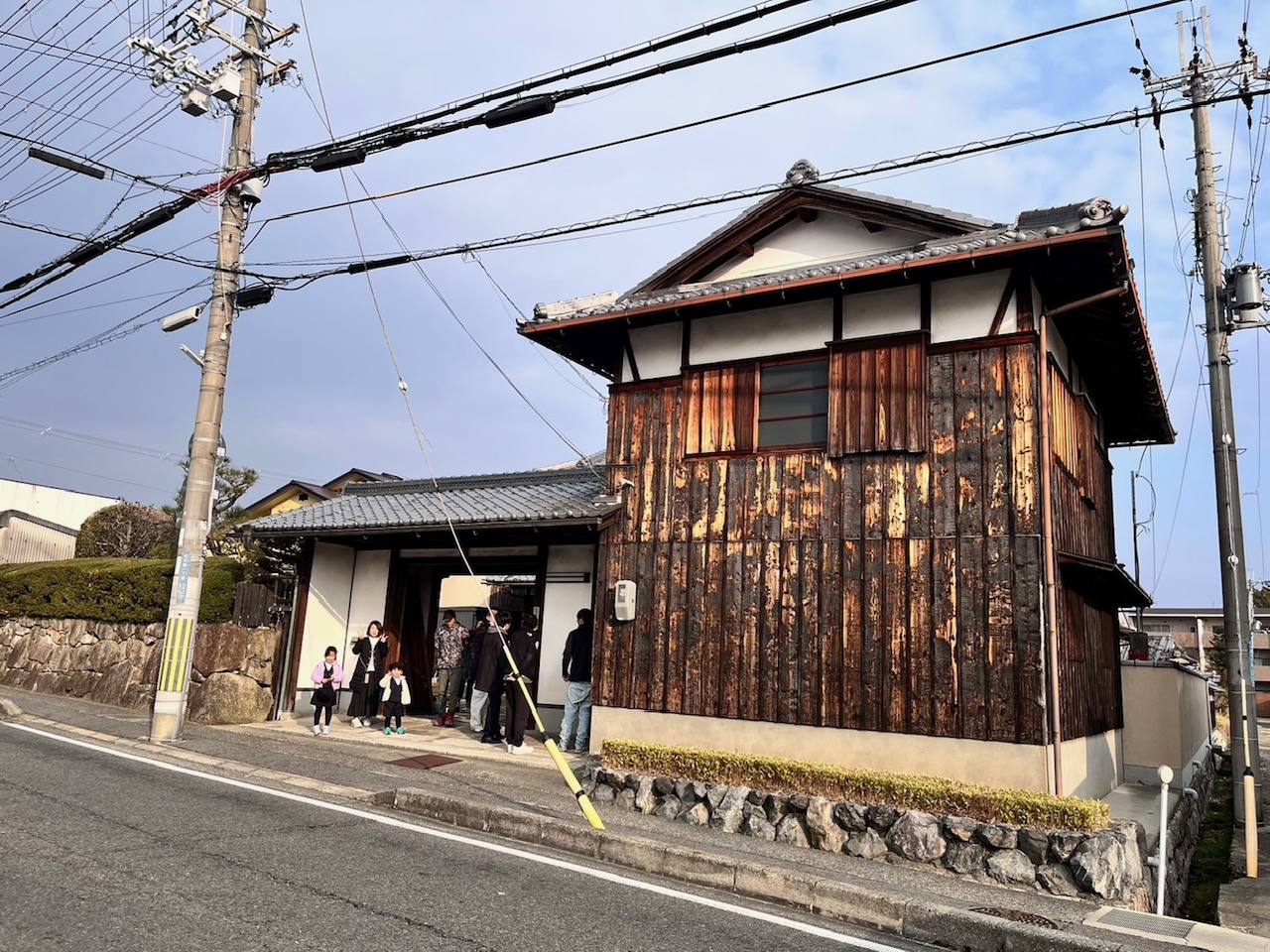 Group of people and children standing outside a weathered two-story wooden Japanese house on a street with utility poles and wires.
