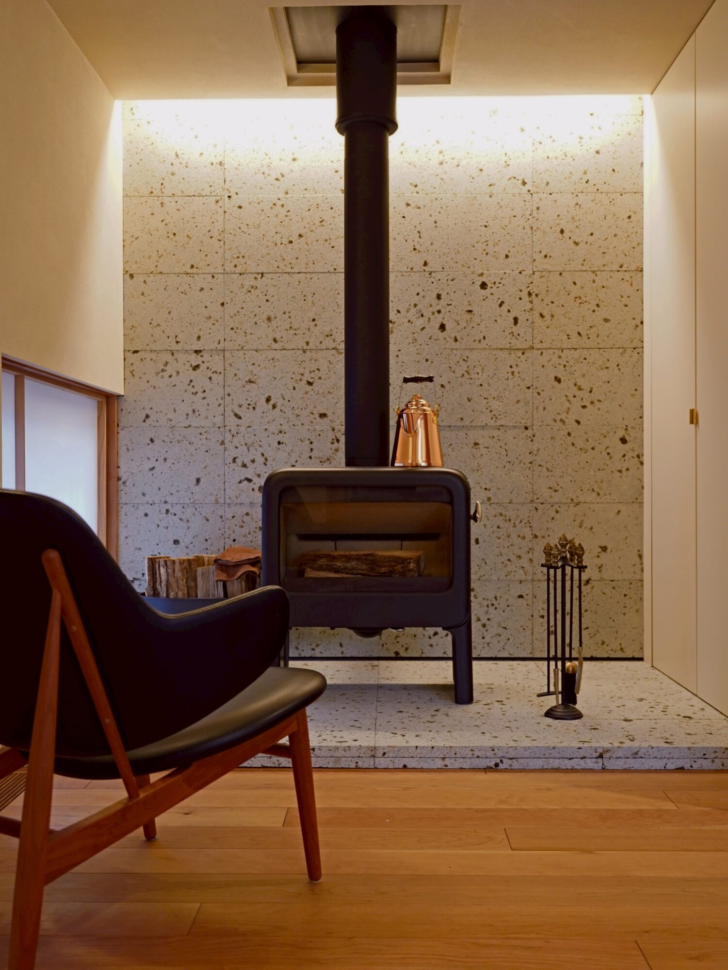 Wood-burning stove centered in a living room with a black flue pipe, copper kettle on top, and fireplace tools to the right; a chair in the foreground.