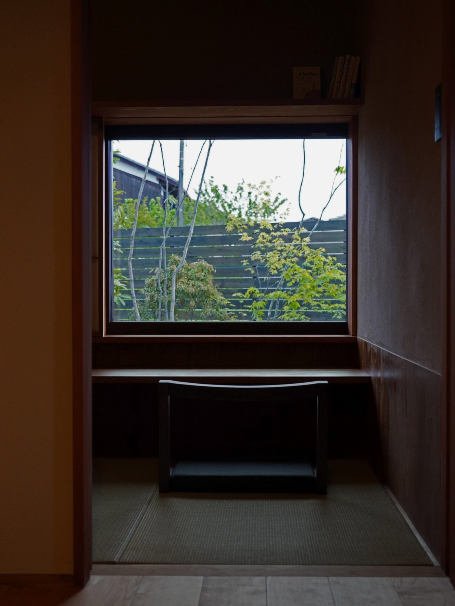 Dimly lit room with a square wooden-framed window opening to a garden with green plants and a wooden fence; a dark chair sits beneath the window.