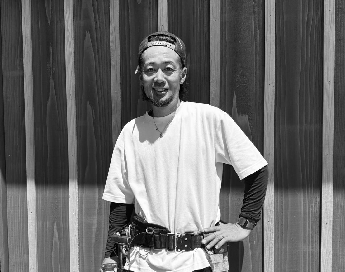 Man wearing a white t-shirt and backward cap, standing with hand on hip in front of wooden siding; tool belt with tools visible at his waist.