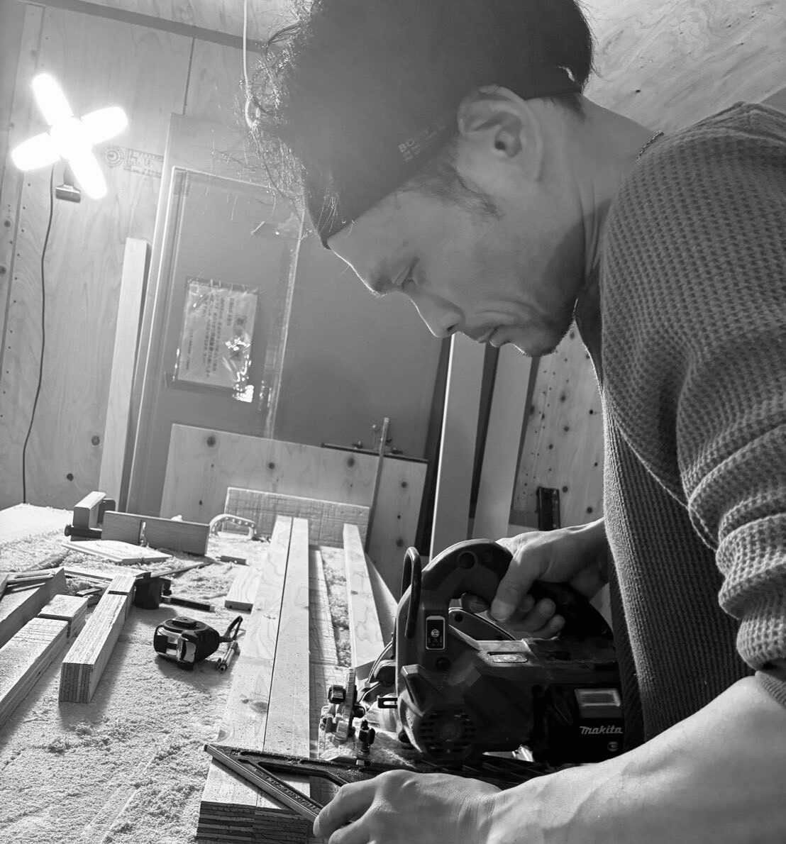 Man using a power saw to cut a wooden plank in a workshop, surrounded by lumber and tools in grayscale.