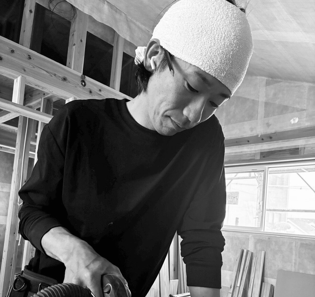 Man wearing a white head wrap and black shirt uses a power sander on a construction site with wooden framing.