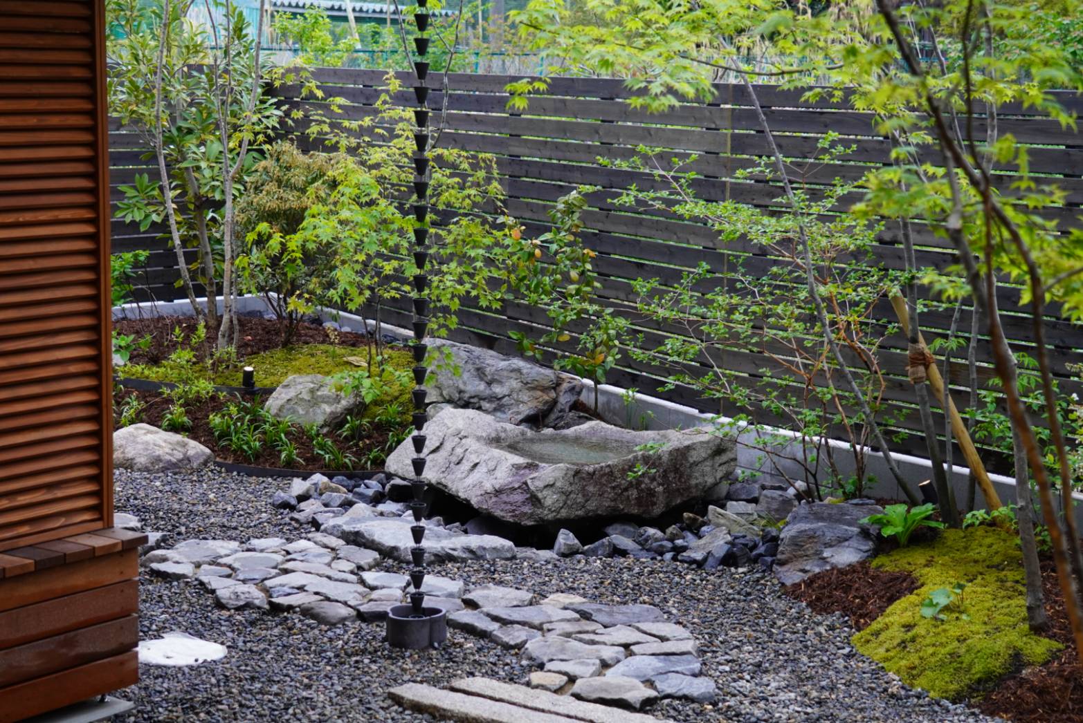 Japanese-style rock garden with a tiered black cup water feature among moss, rocks, and a gravel path beside a wooden-clad structure and dark slatted fence.
