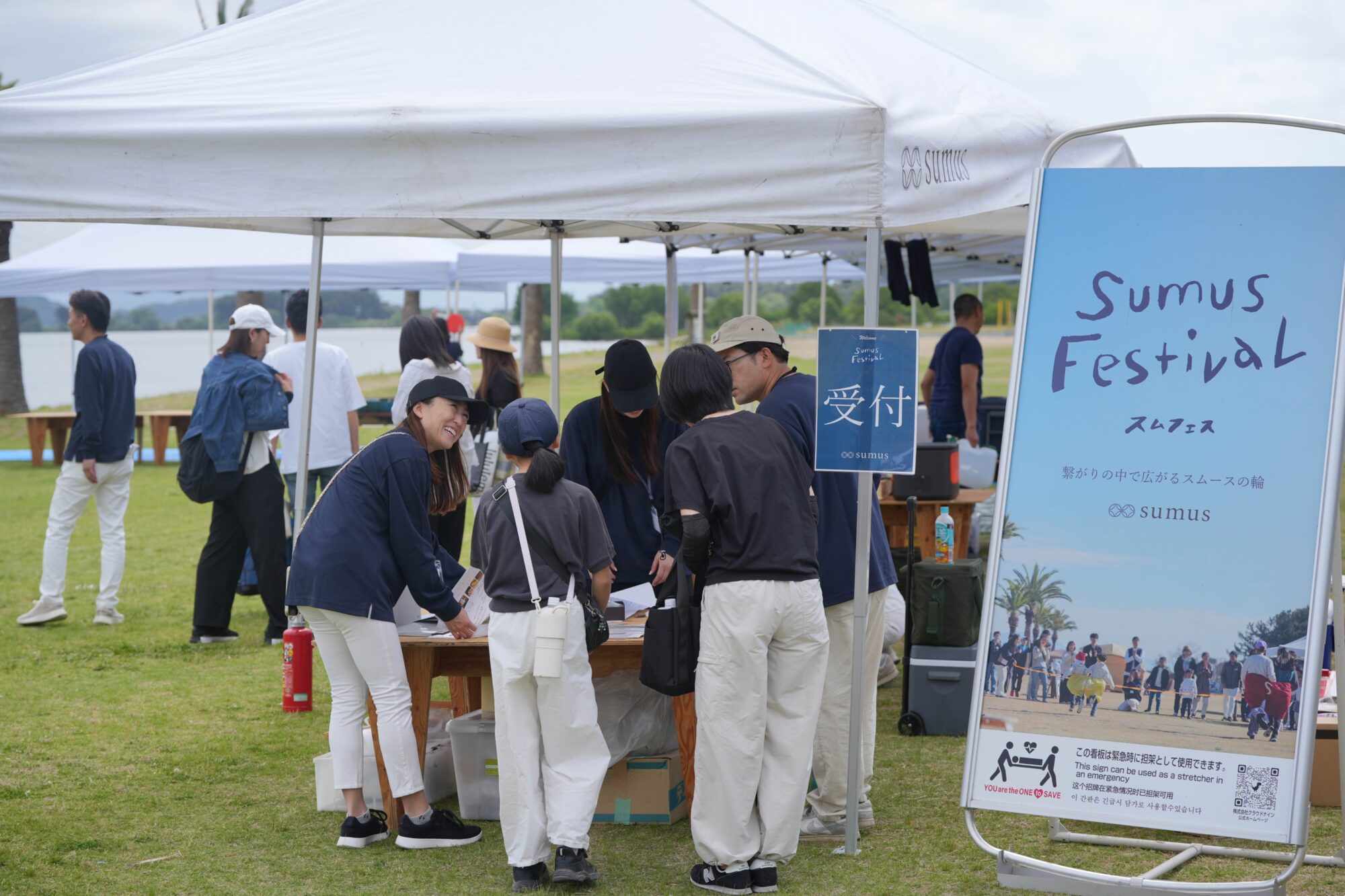 Crowd at an outdoor festival under white tents; a large blue Sumus Festival sign on the right.