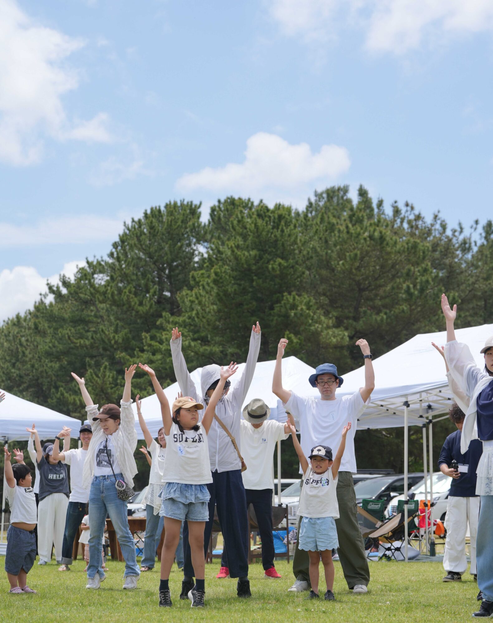Group of people outdoors with arms raised, participating in a communal activity under white tents and a blue sky.
