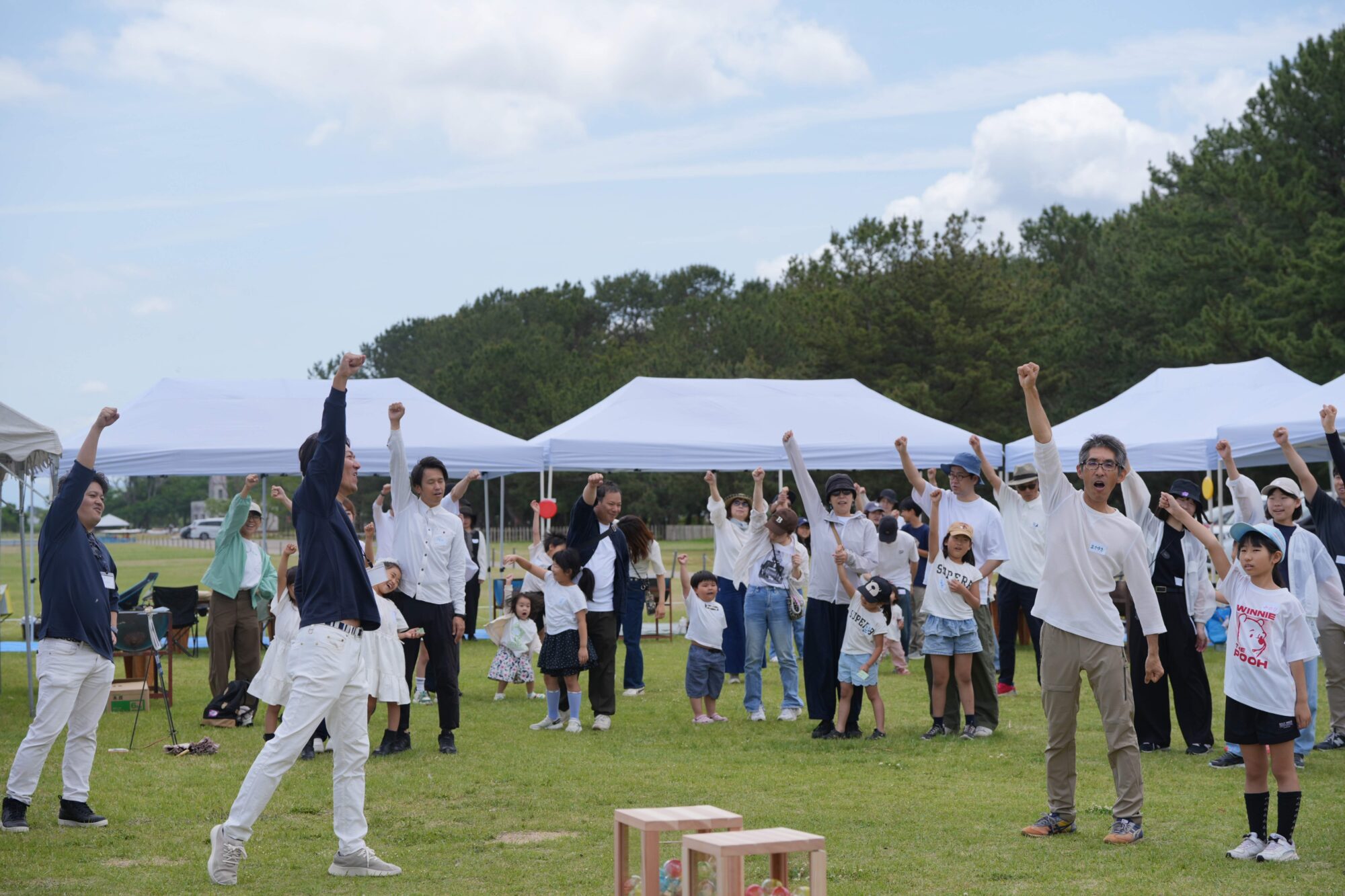 Crowd of people outdoors under white tents raising fists in celebration on a sunny day in a park with trees in the background