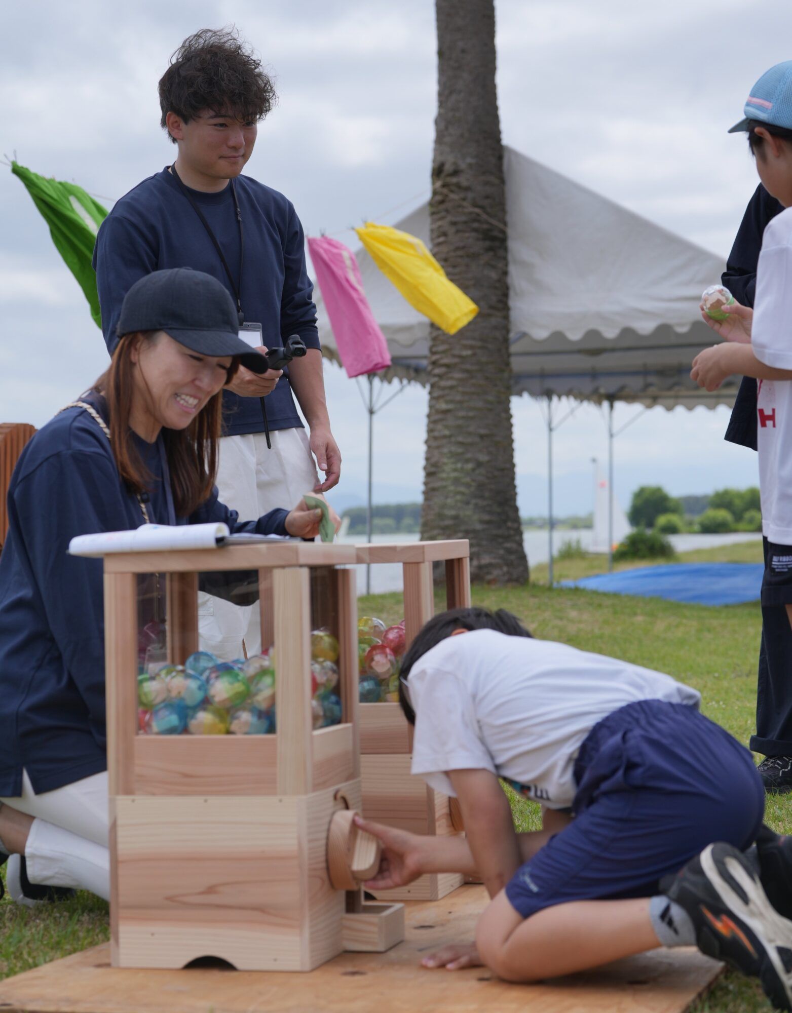 Young man with a lanyard beside a wooden ball-dispensing box filled with colorful marbles at an outdoor festival or event.