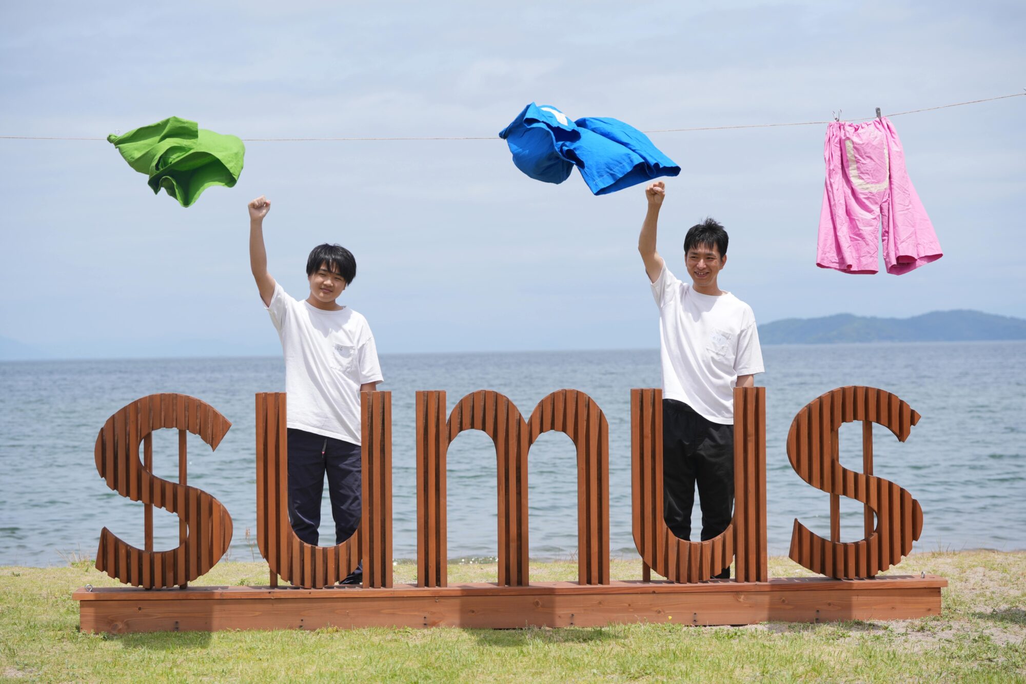 Two men at a beach raise their fists behind a large rust-colored wooden sign with curved letters, with clothes hanging on a line above and the ocean and sky in the background.