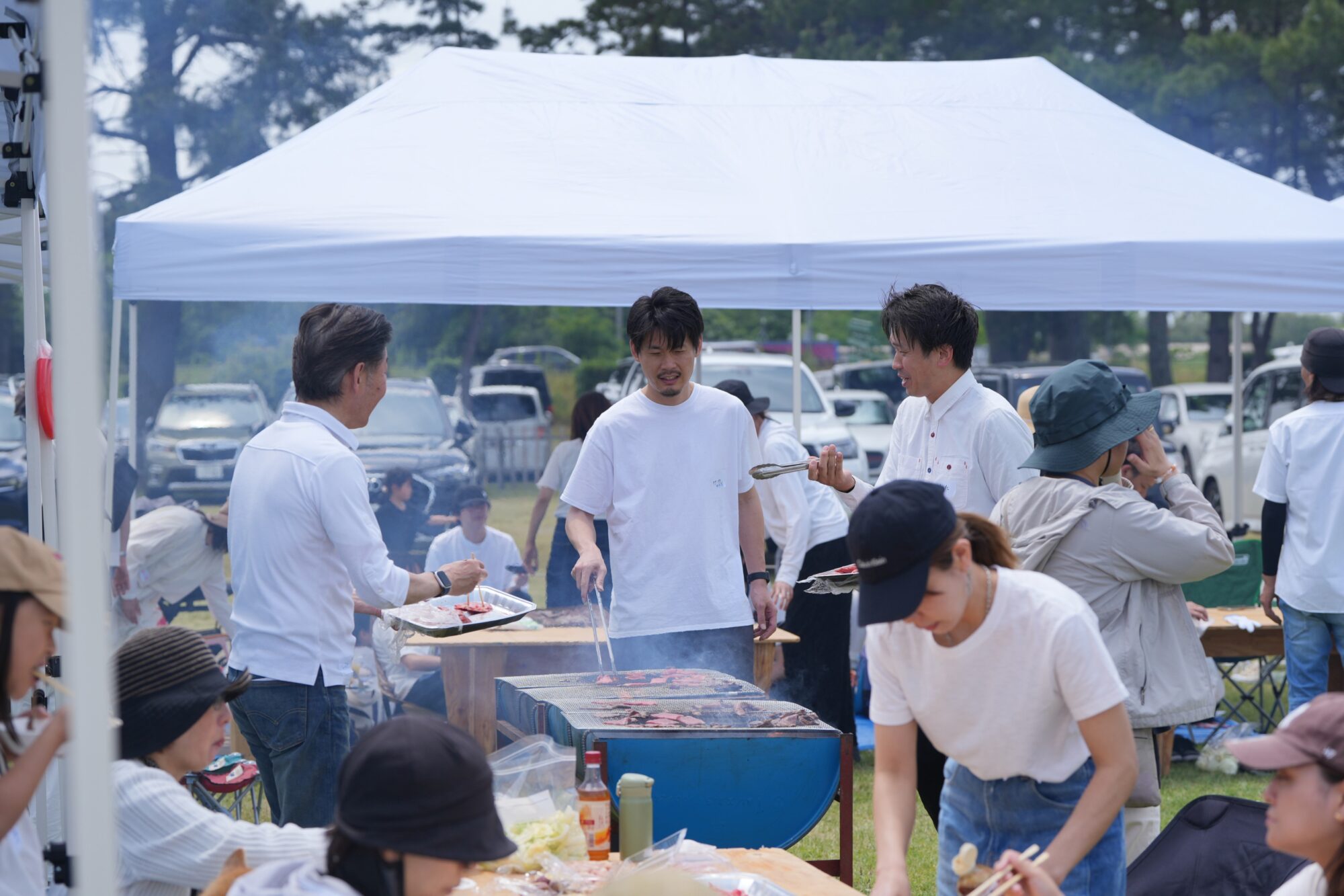 Group of people at an outdoor barbecue under a white canopy, grilling meat and serving food under a sunny park.
