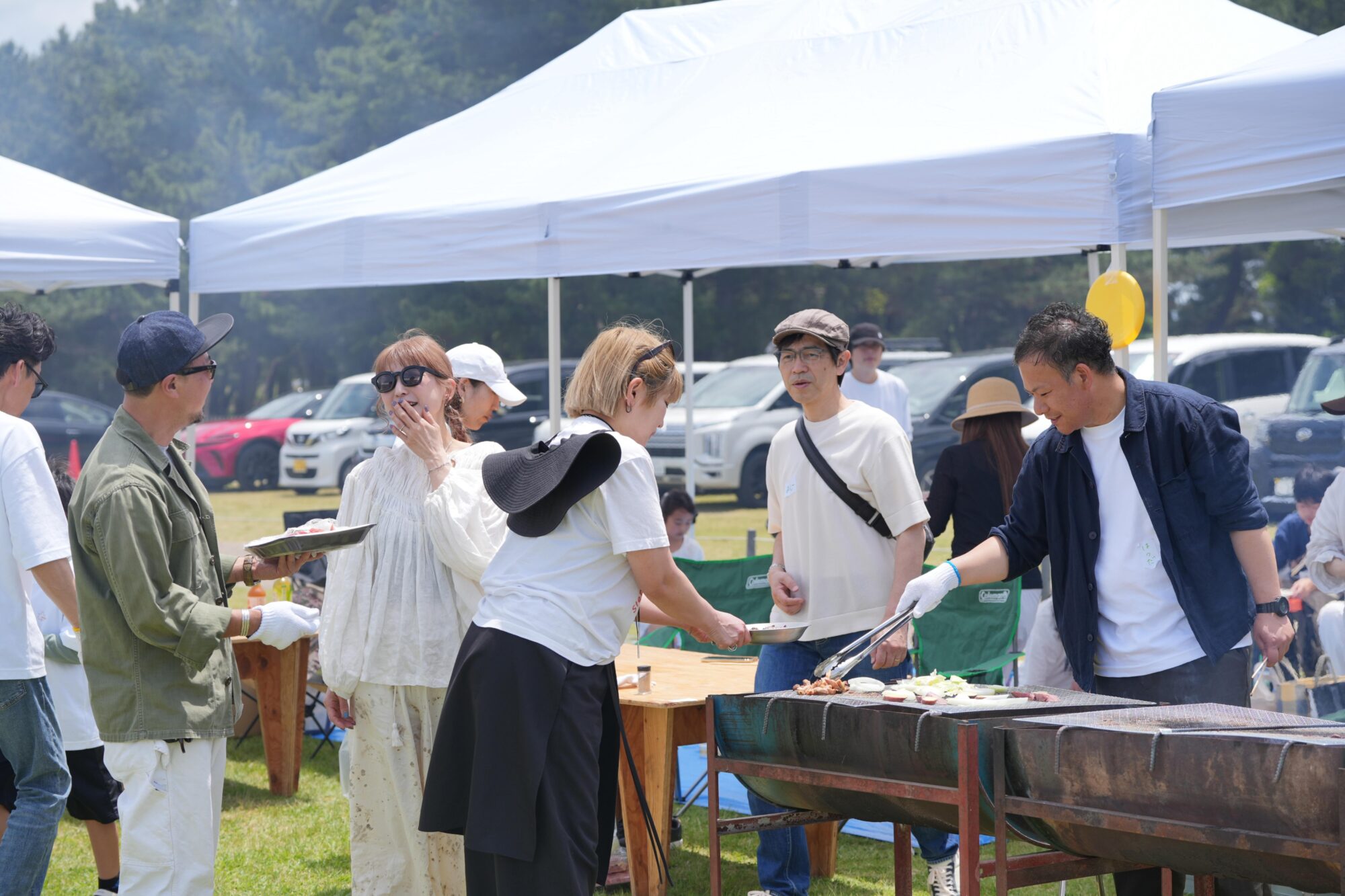 People at an outdoor barbecue under white canopy tents, grilling and serving food to guests on a sunny day.