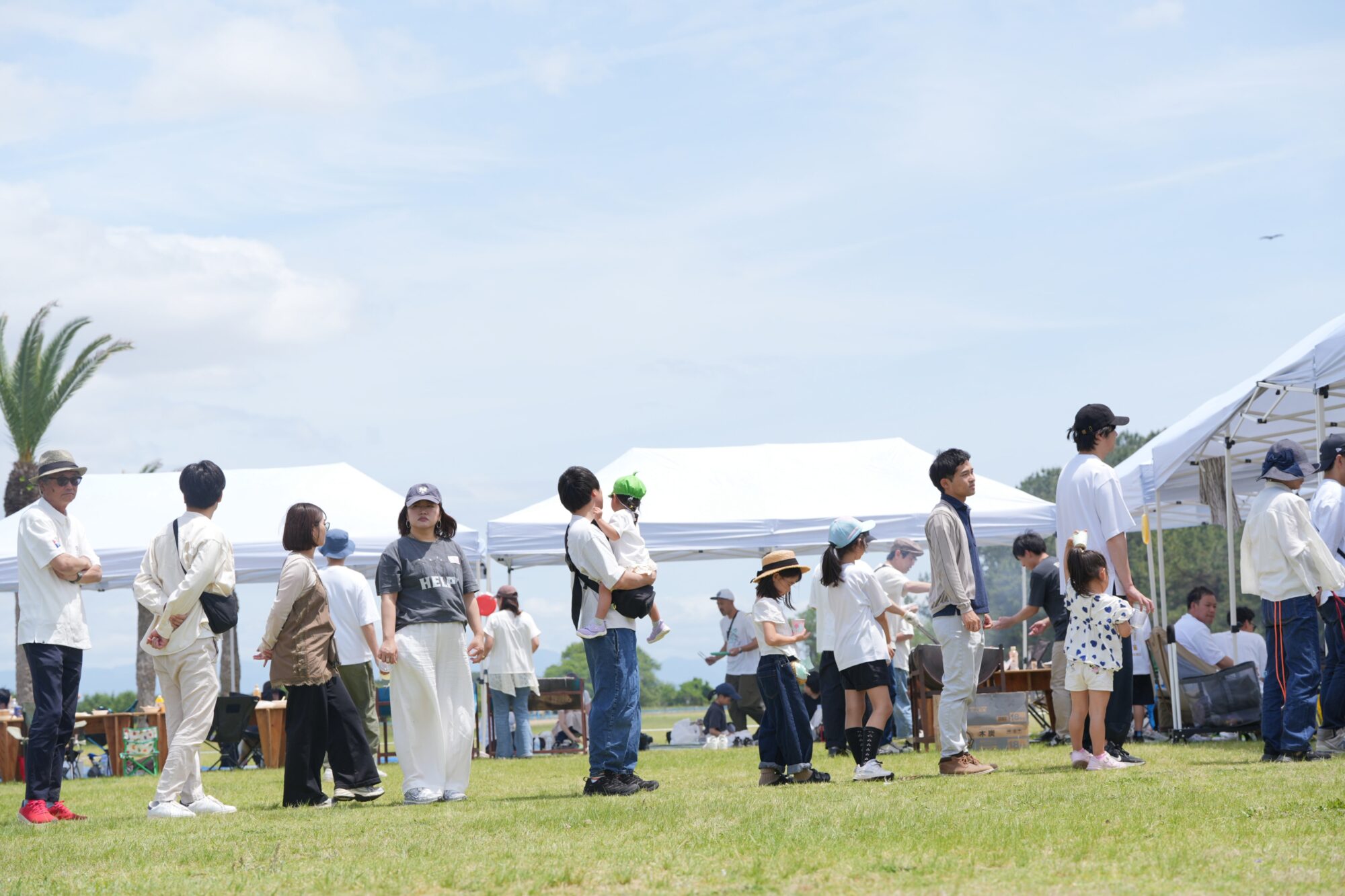 Group of people standing and walking on a grassy field beside white tents under a bright blue sky at an outdoor event.