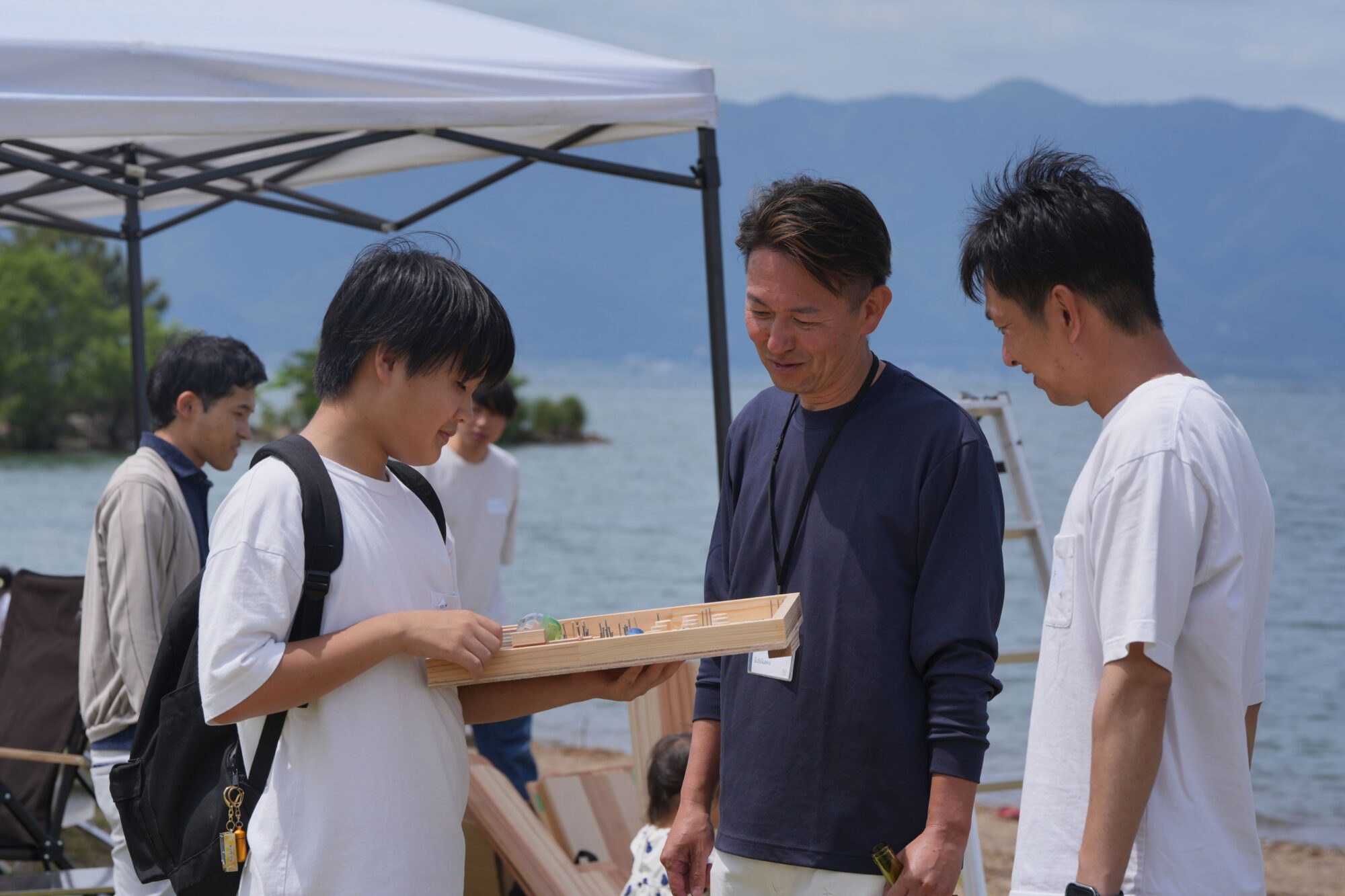 Group of people outdoors by a lake, inspecting a wooden board/puzzle under a canopy with mountains in the background, smiling.