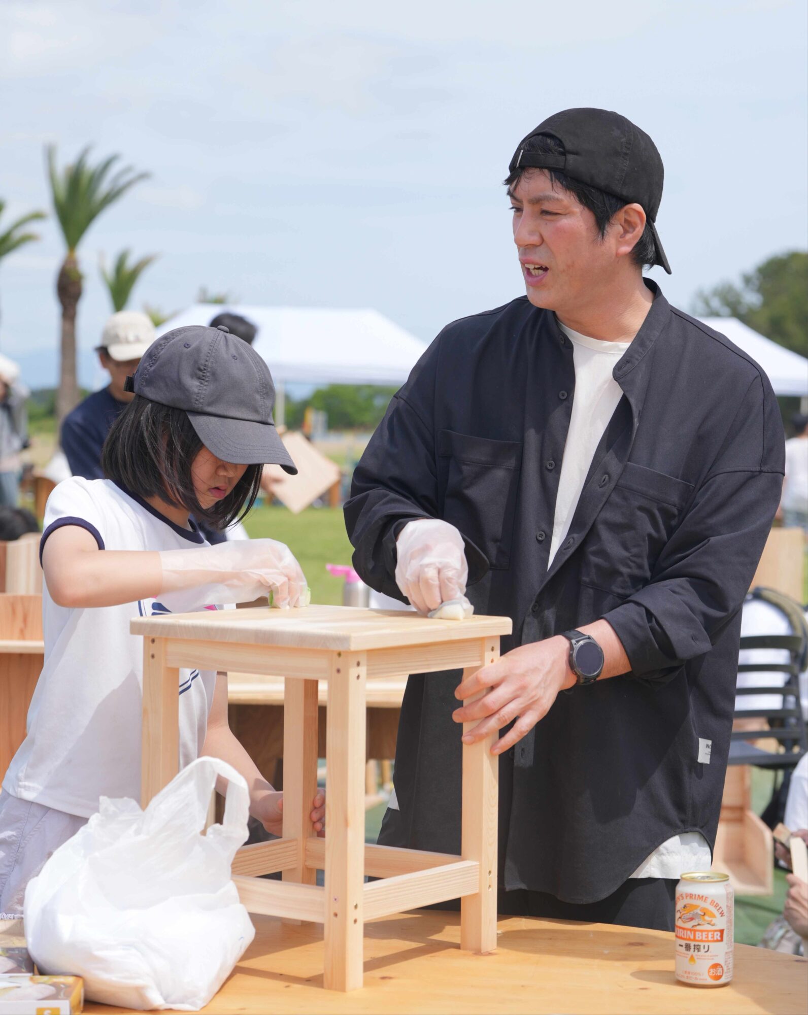 Man wearing a backward black cap assists a young girl with a wooden woodworking project at an outdoor booth; a beverage can sits on the table nearby.
