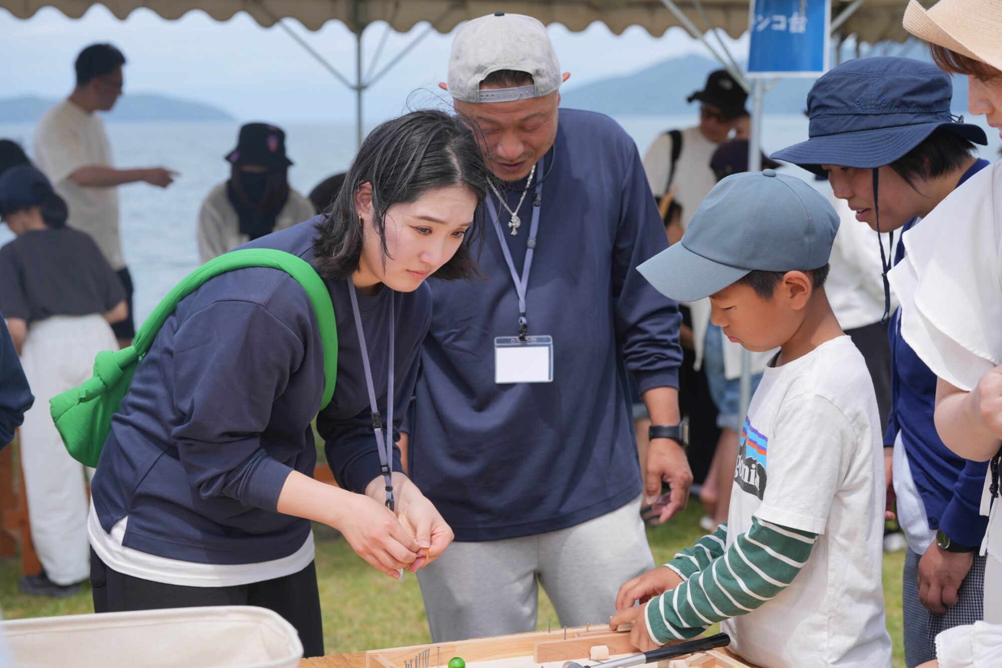 Group of people at a lakeside outdoor event, focused on a wooden stick game on a table.