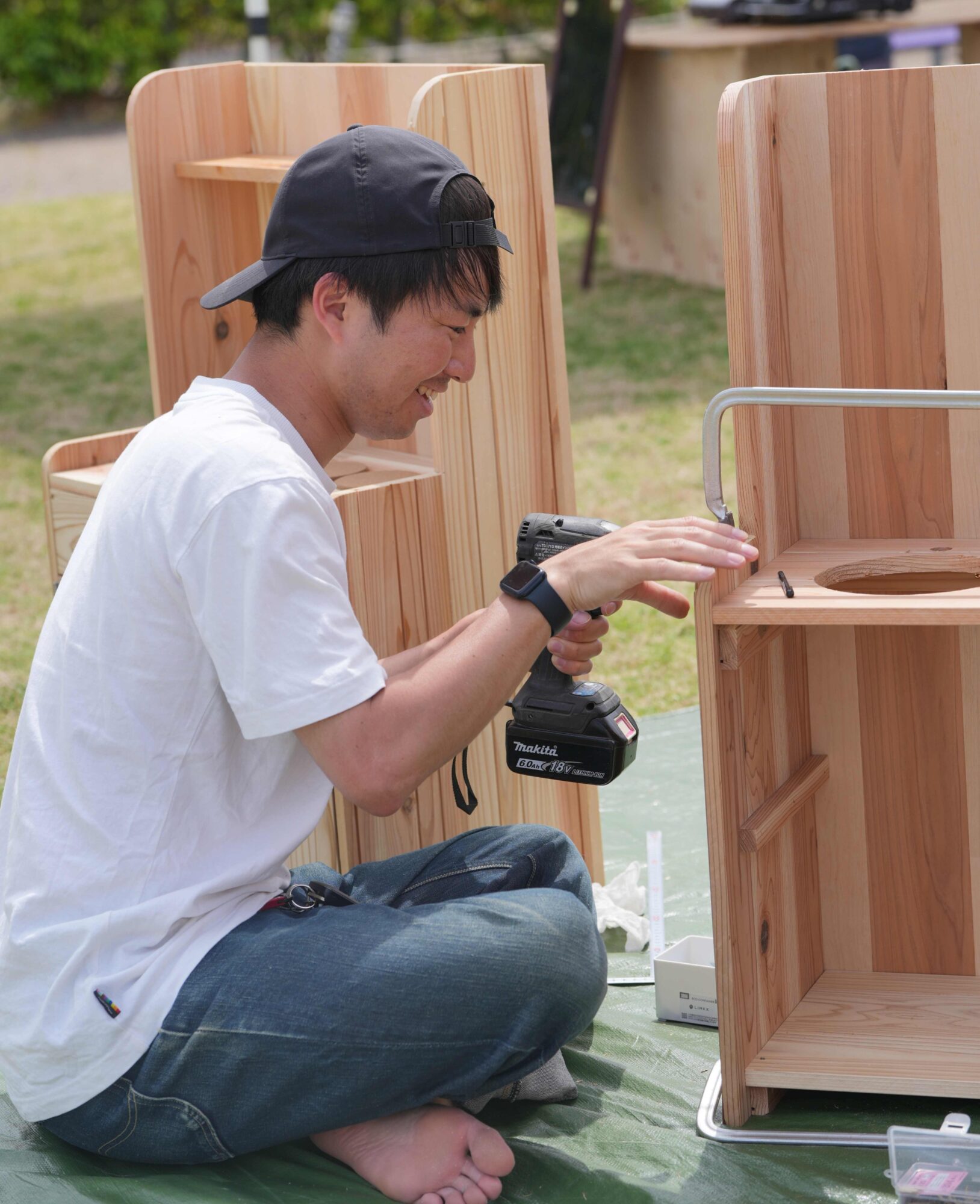 Man kneeling outdoors, wearing a white shirt and cap, using a power drill to assemble tall wooden panels.