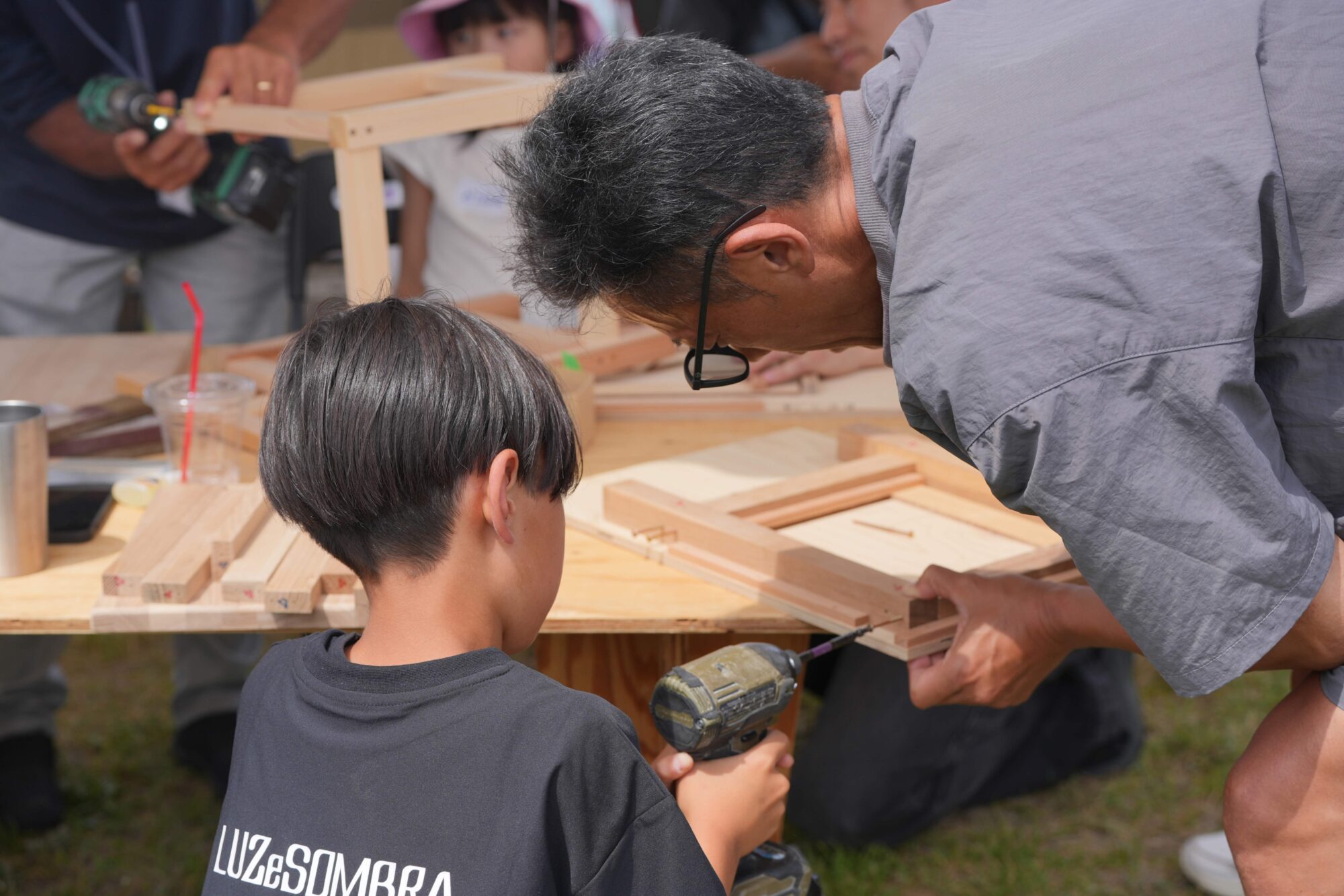 Man with glasses bending over to guide a child using a cordless drill on a wooden frame outdoors at a workshop.