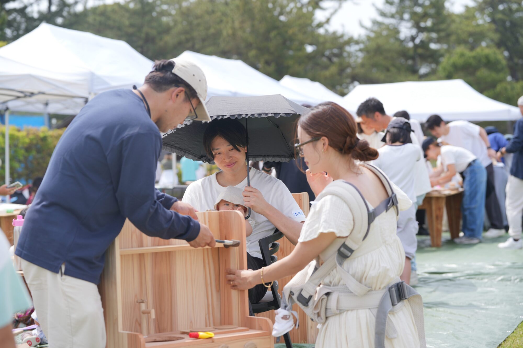 Man in a blue shirt and beige cap helps a child with a wooden craft at an outdoor booth under a parasol, with people and white tents in the background at a festival.
