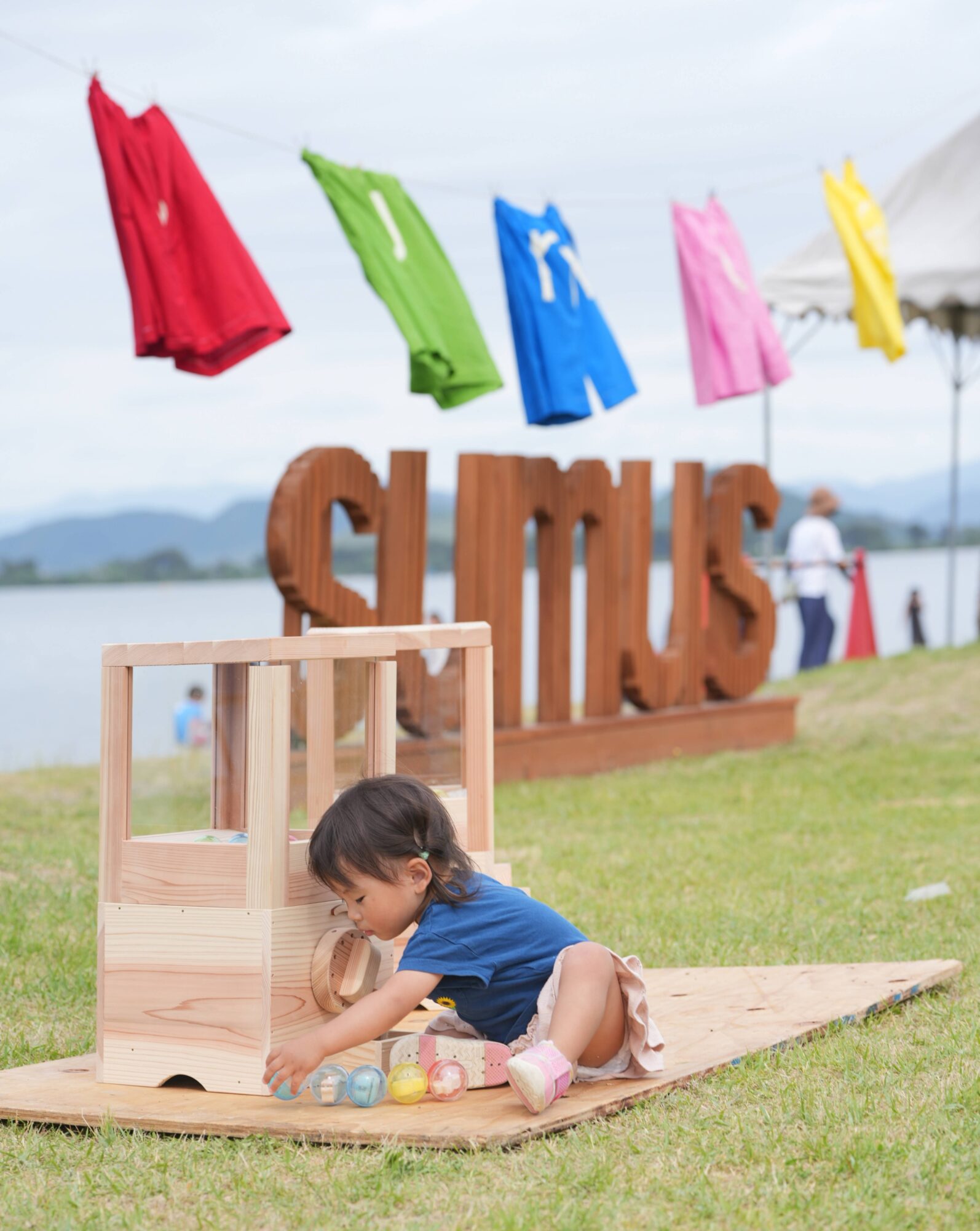 Child in a blue shirt playing with a wooden toy on a grassy field with colorful flags in the background, outdoors.