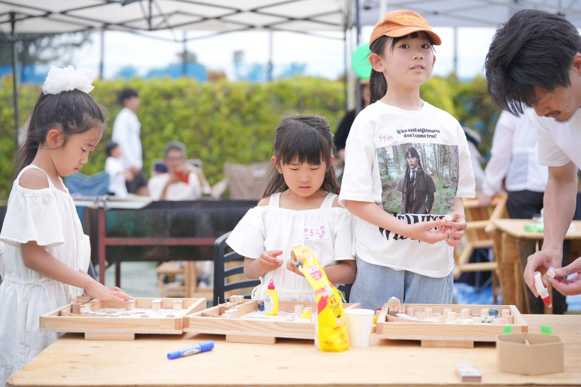 Children participate in an outdoor crafts activity at a wooden table under tents, using glue and wood pieces.