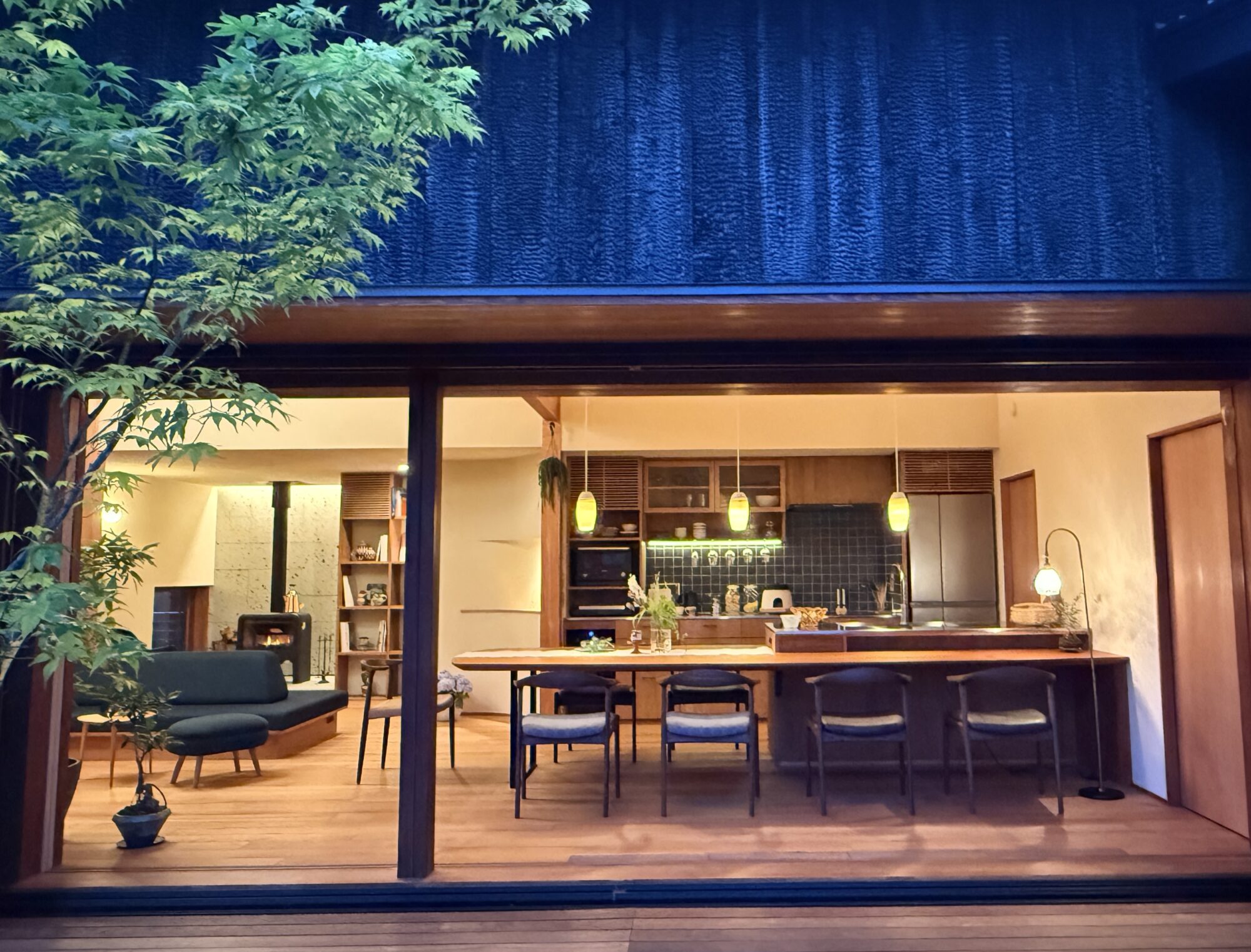 Cafe interior with wooden tables and chairs, left-side potted plant, and a blue textured wall behind an open kitchen area with pendant lights.