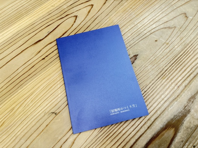 Blue booklet lying on a light wooden table, with small white Japanese text on the lower-right corner of the cover