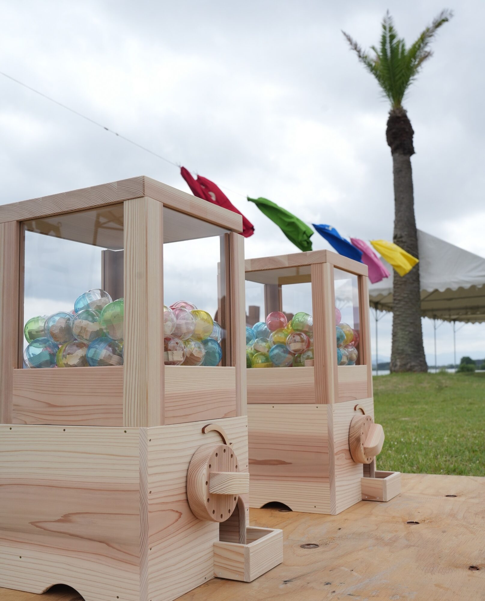 Wooden toy train car filled with translucent colorful balls outdoors, with rainbow flags and a palm tree in the background.