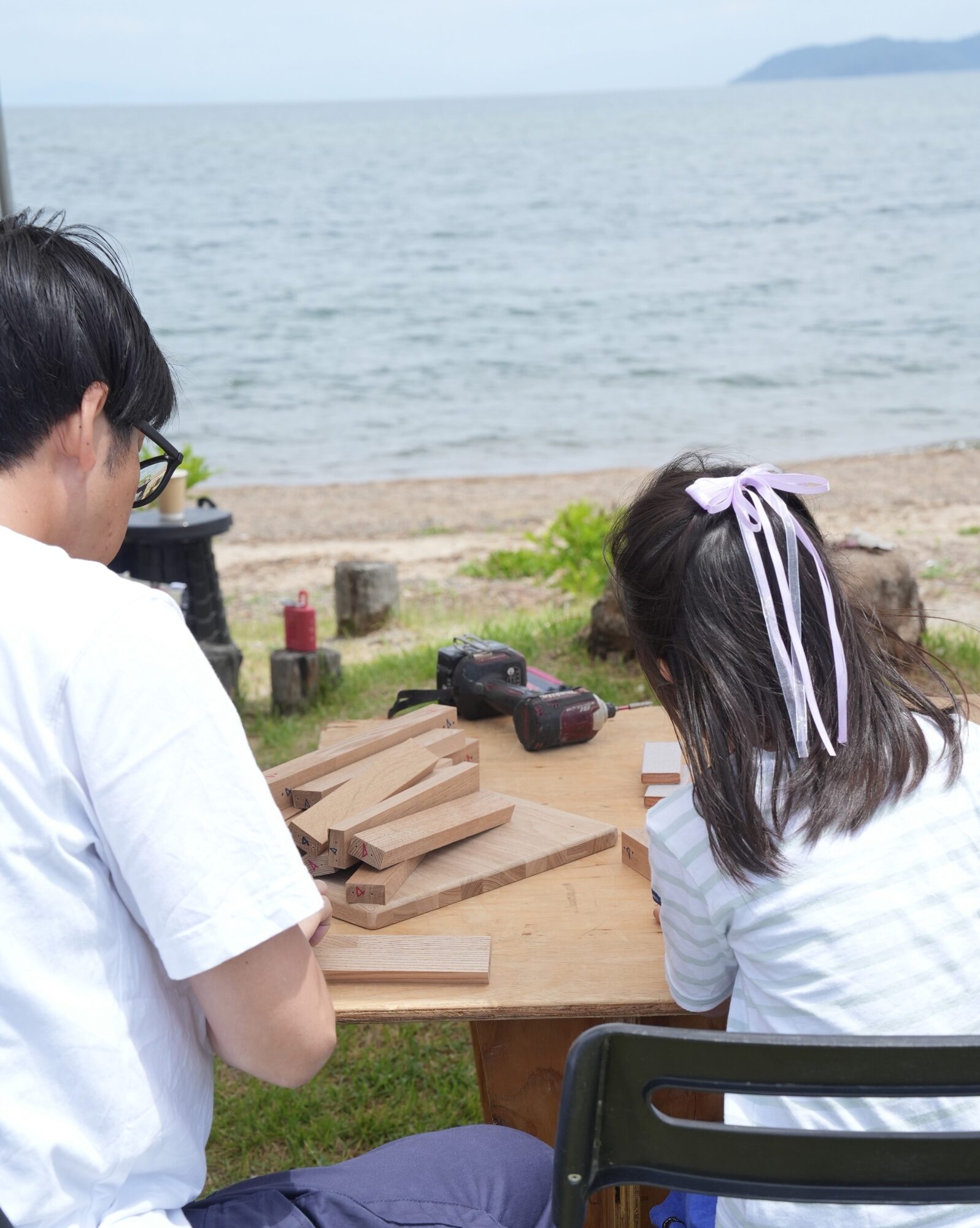 Two people at a beach-side work table arranging wooden blocks, with a drill and tools nearby and the ocean in the background.