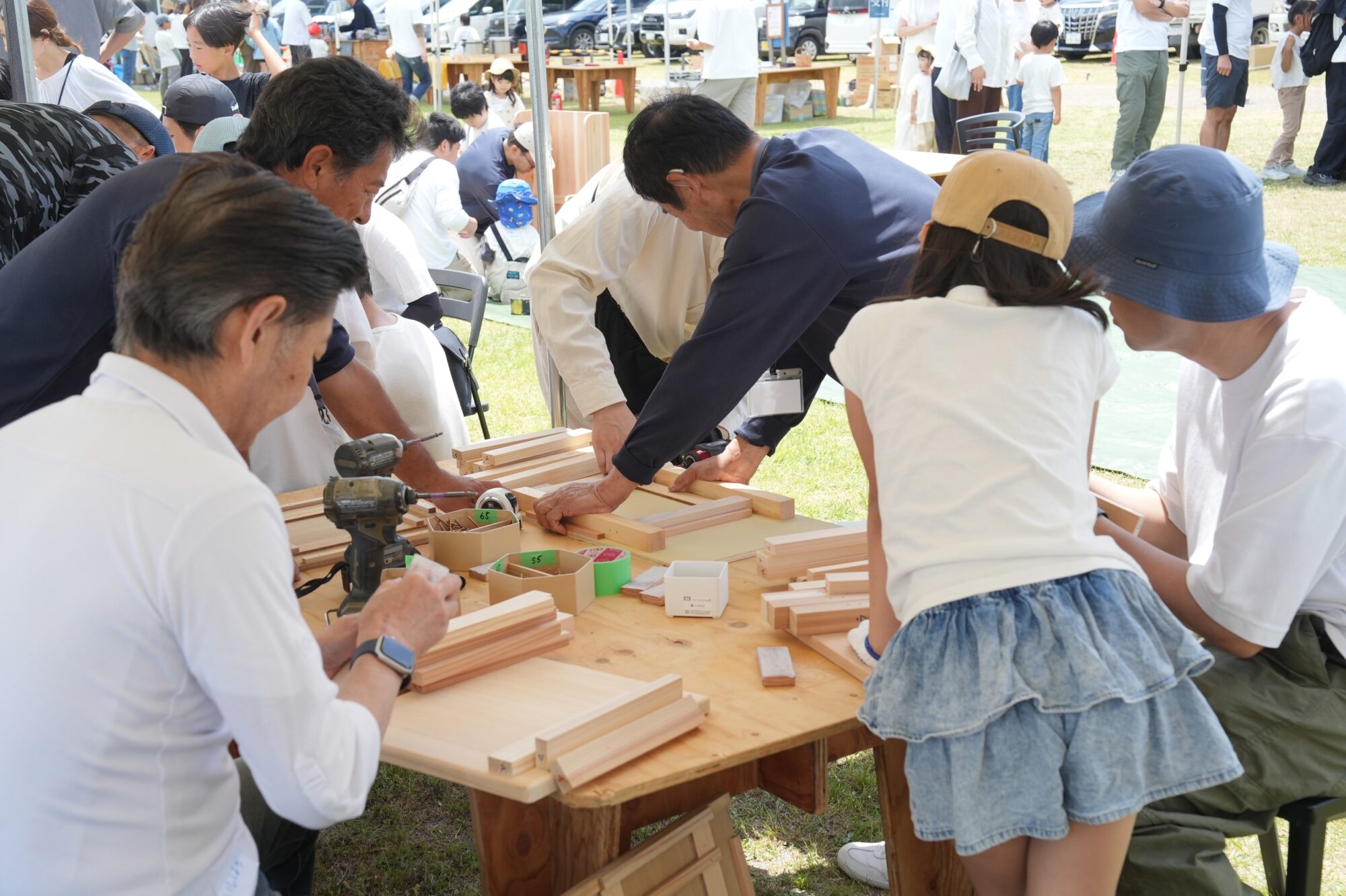 People gathered around a wooden workbench outdoors, using tools to cut and assemble slim wooden boards.