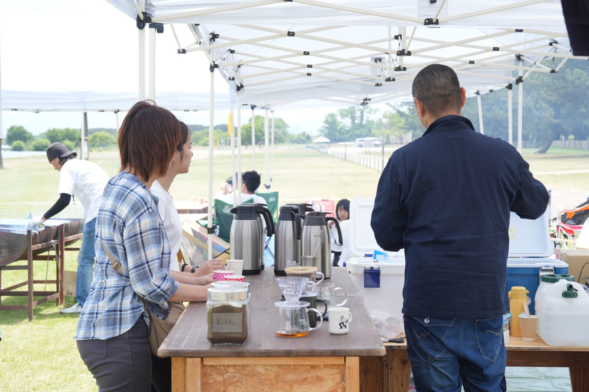 Woman in a plaid shirt and a man in a blue jacket serve coffee at an outdoor booth under white canopy tents; a grassy field is in the background.