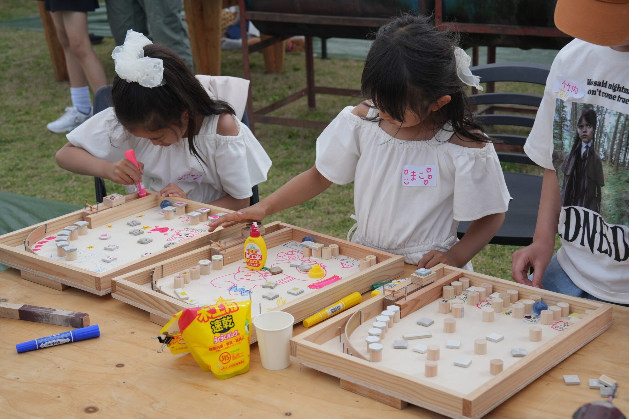 Two children in white tops work on a craft project at an outdoor wooden table filled with small wooden tiles, markers, and glue.