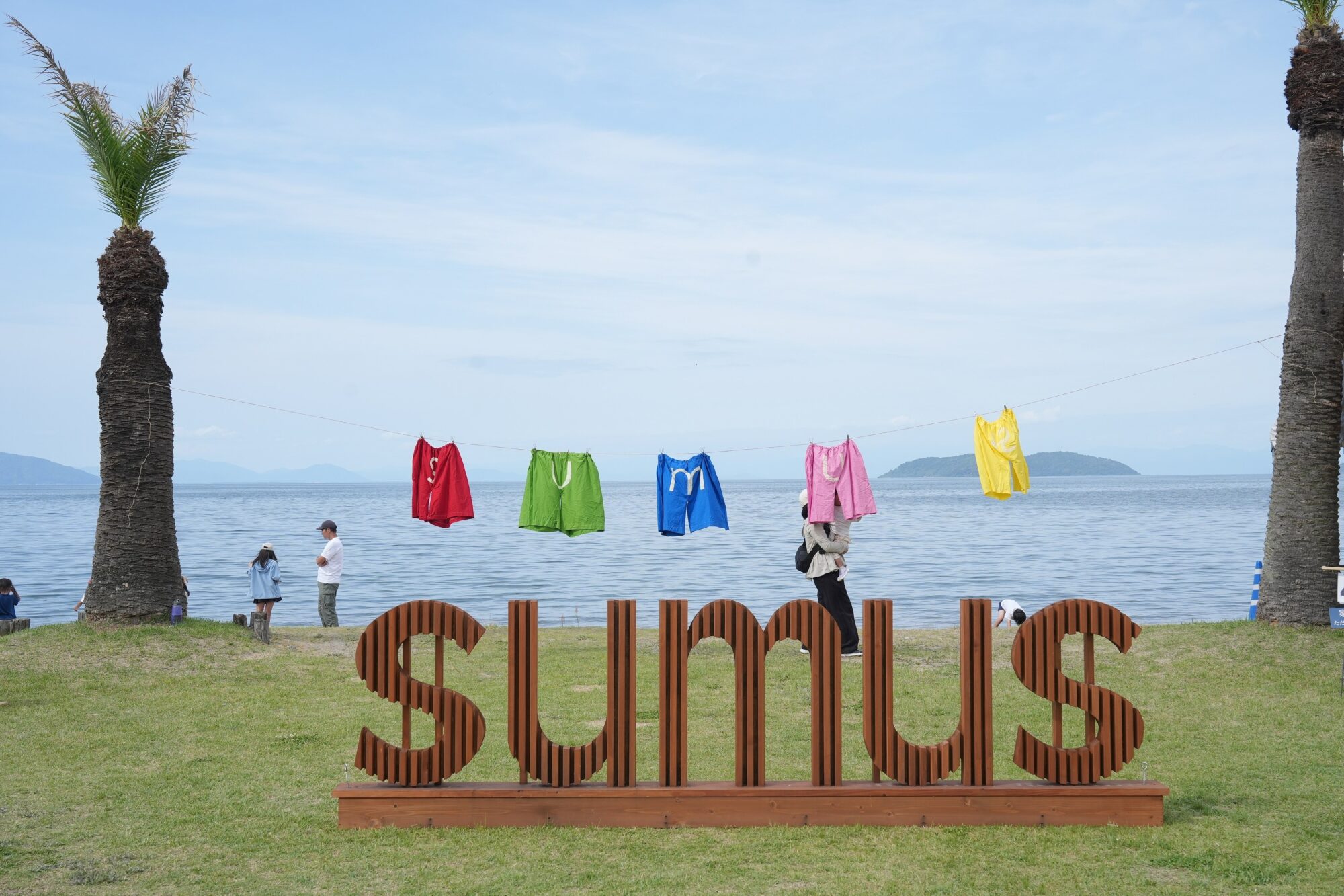 Colorful clothes hang on a line by the sea, with two palm trees and a large brown sculpted word 'Sea' in the foreground. A calm beach and distant islands are in the background.