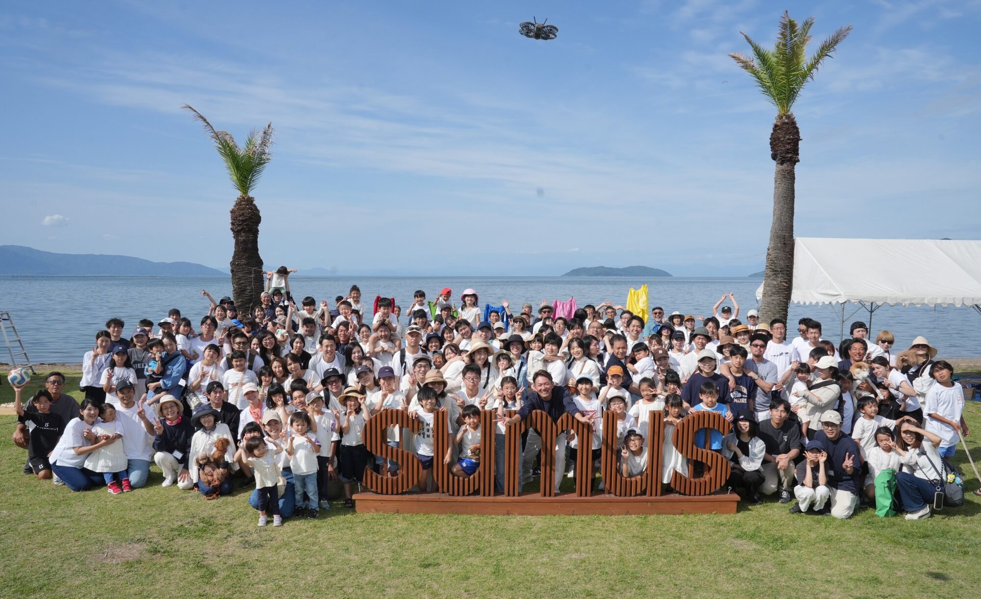 Large group of people posing for a photo on a grassy area by the sea, with palm trees, a white tent, and a drone in the blue sky above.