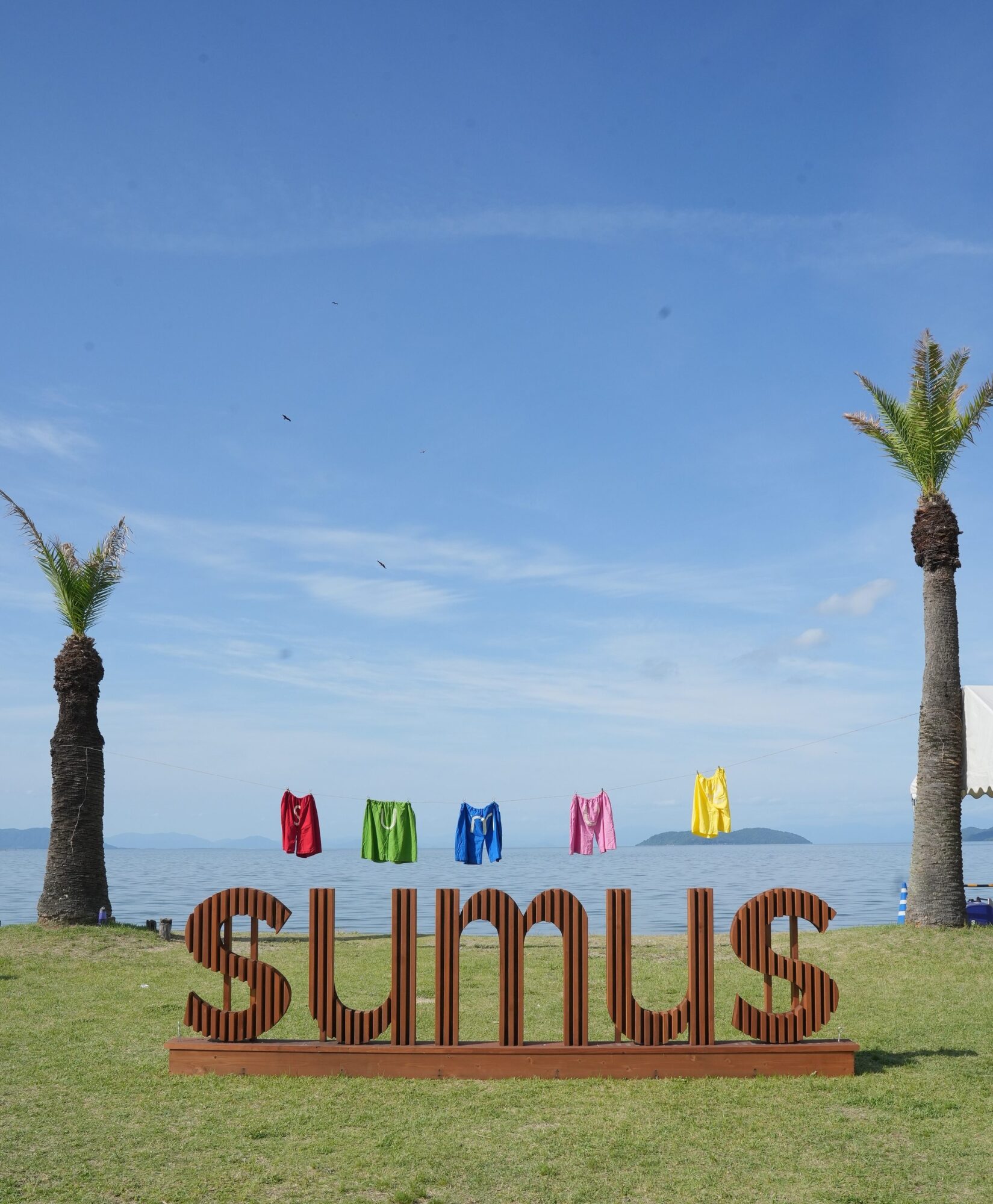 Coastal park scene with palm trees, a line of colorful clothes hanging to dry, and a large brown word sculpture by the water. The sky is blue and calm sea is in the background.