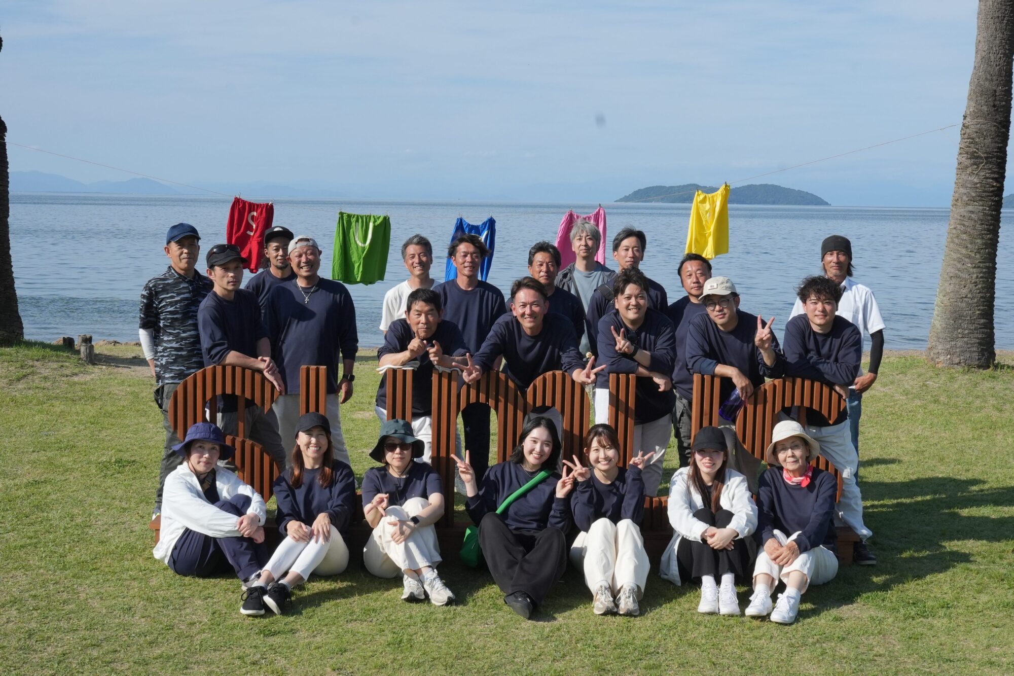 Group of people posing for a photo on a grassy shore by a calm lake, with colorful clothes hanging on a line behind them and an island in the distance.