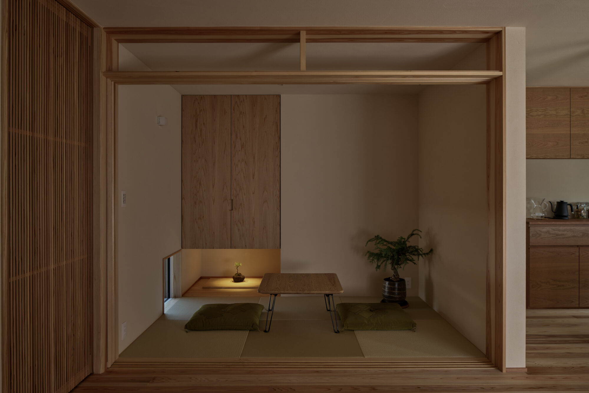 260217_019 - 滋賀で自然素材・木の家の注文住宅を建てる工務店|株式会社スムース Japanese-inspired alcove with a low wooden table, tatami mats, cushions, and a potted plant under warm lighting