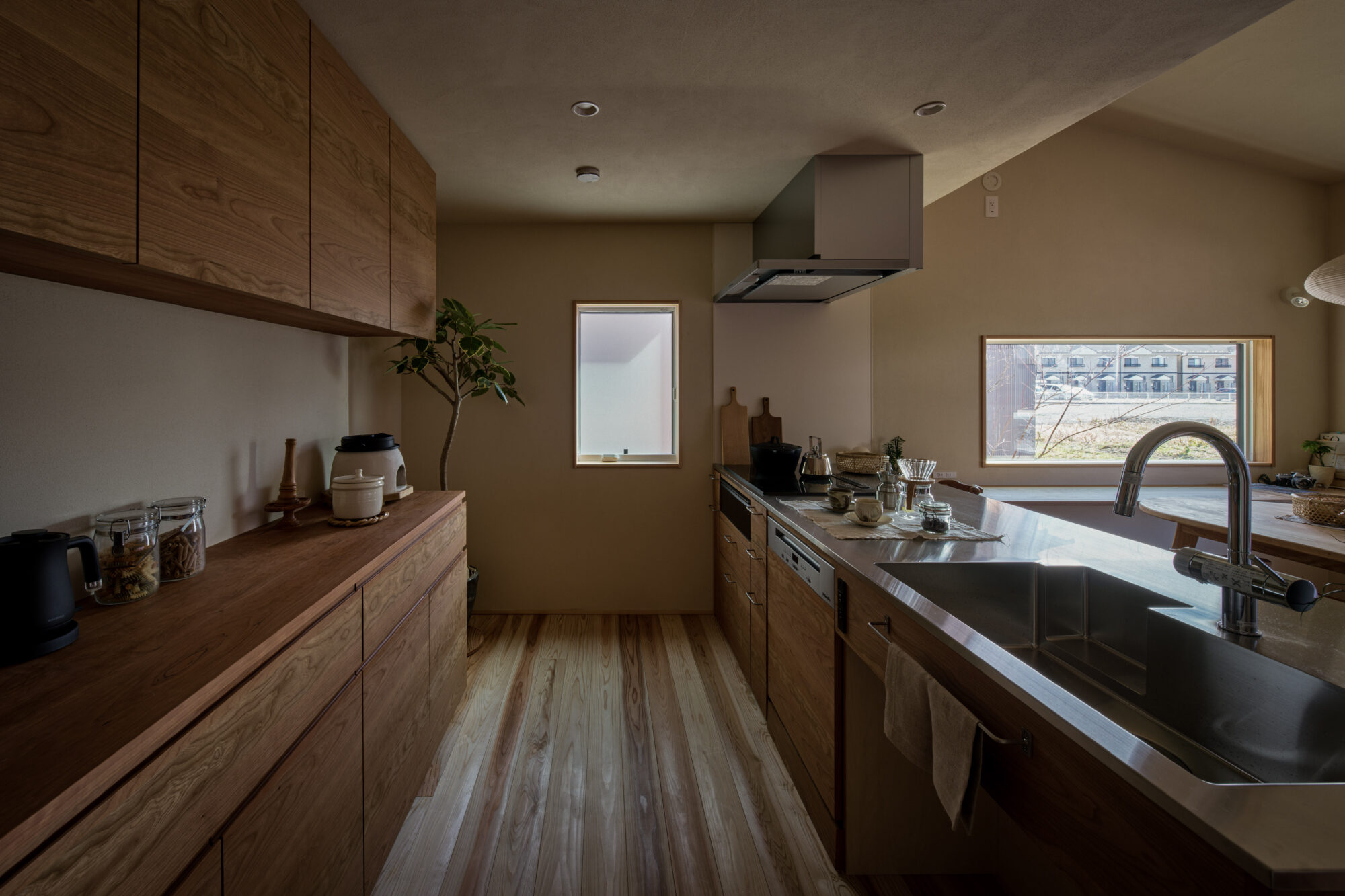 260217_031 - 滋賀で自然素材・木の家の注文住宅を建てる工務店|株式会社スムース Wooden kitchen with long cabinets, stainless steel island, and a double sink under a window looking out at houses outside; warm natural light and a potted plant on the counter.