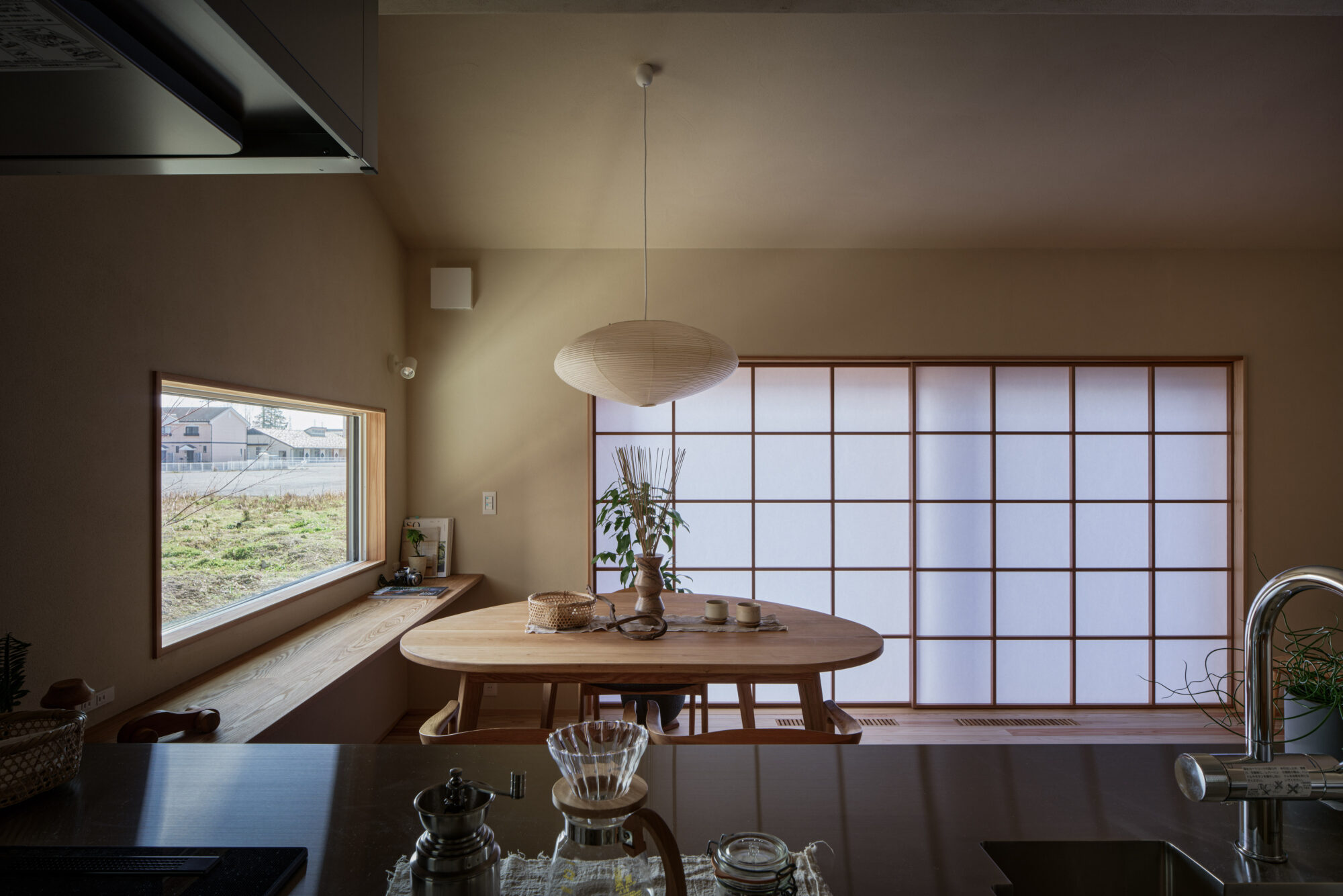 260217_033 - 滋賀で自然素材・木の家の注文住宅を建てる工務店|株式会社スムース Round paper lantern pendant lamp hangs over a wooden dining table in a warm, minimalist room with shoji screens and a window bench.