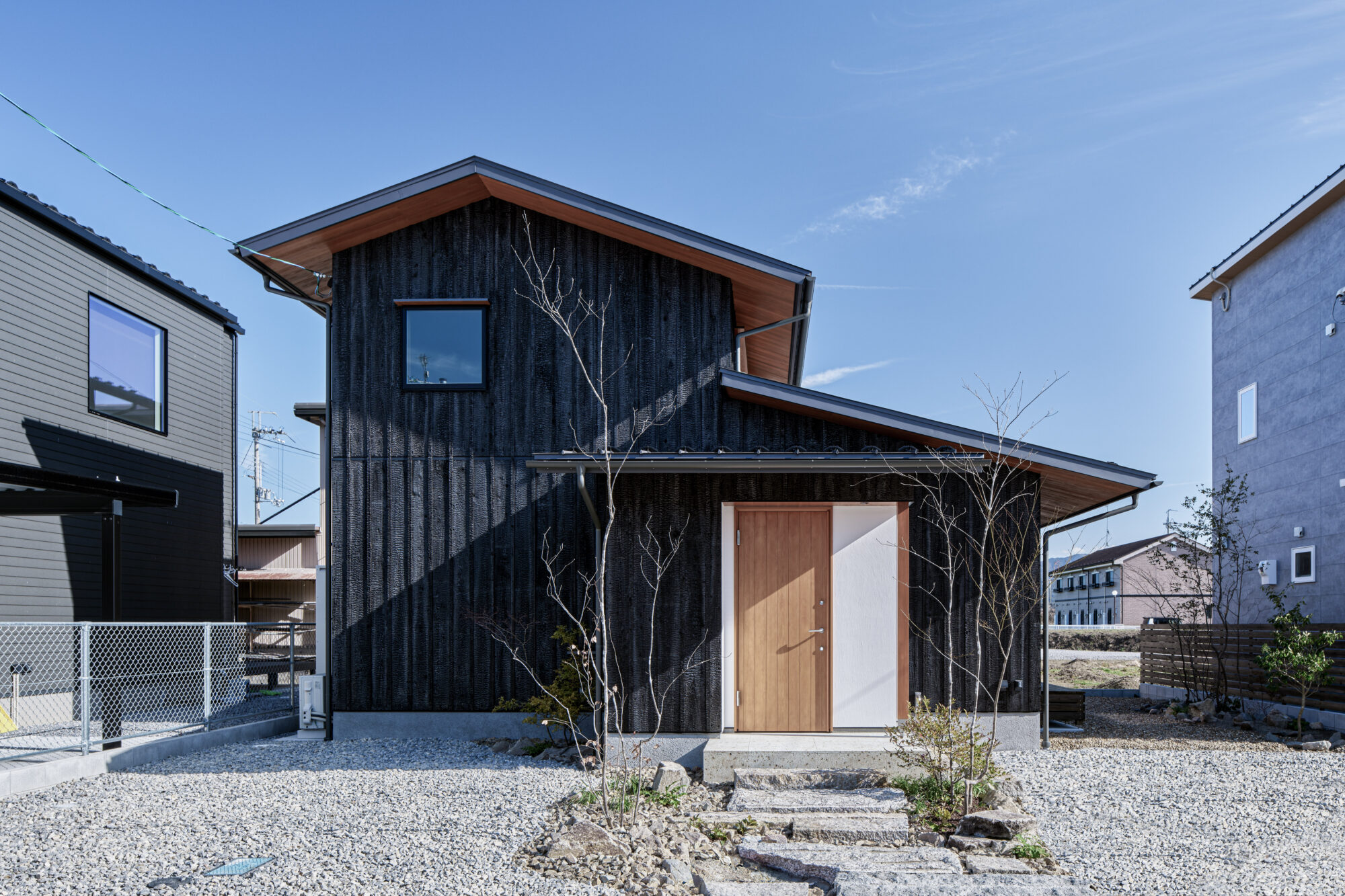 260217_070 - 滋賀で自然素材・木の家の注文住宅を建てる工務店|株式会社スムース Front view of a modern house with black vertical siding, a wooden front door, and a gabled roof; gravel yard with leafless trees in the foreground.