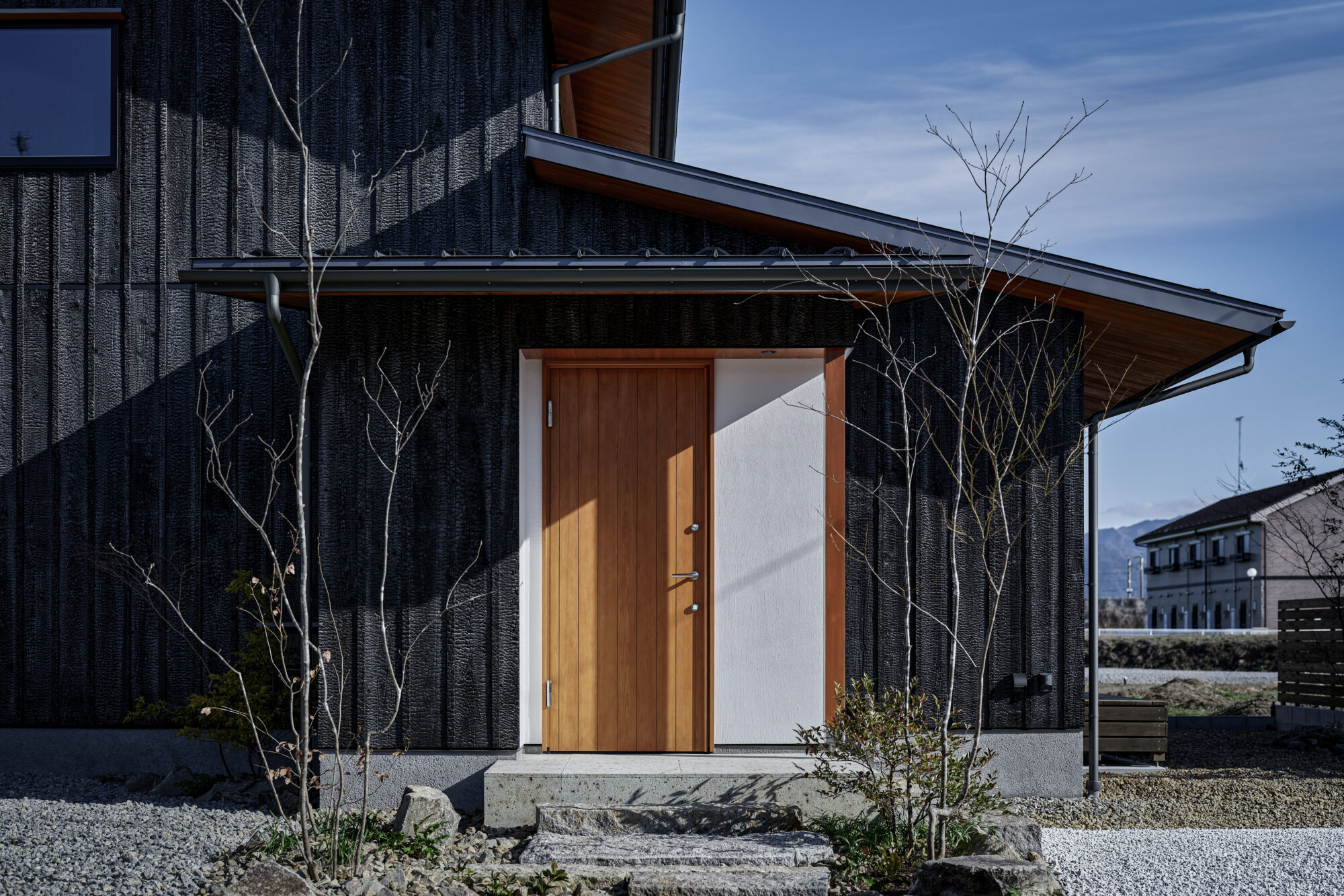 260217_075 - 滋賀で自然素材・木の家の注文住宅を建てる工務店|株式会社スムース Modern house entrance with a brown wooden door set in light concrete, dark textured exterior walls, and leafless trees in a gravel yard under a blue sky