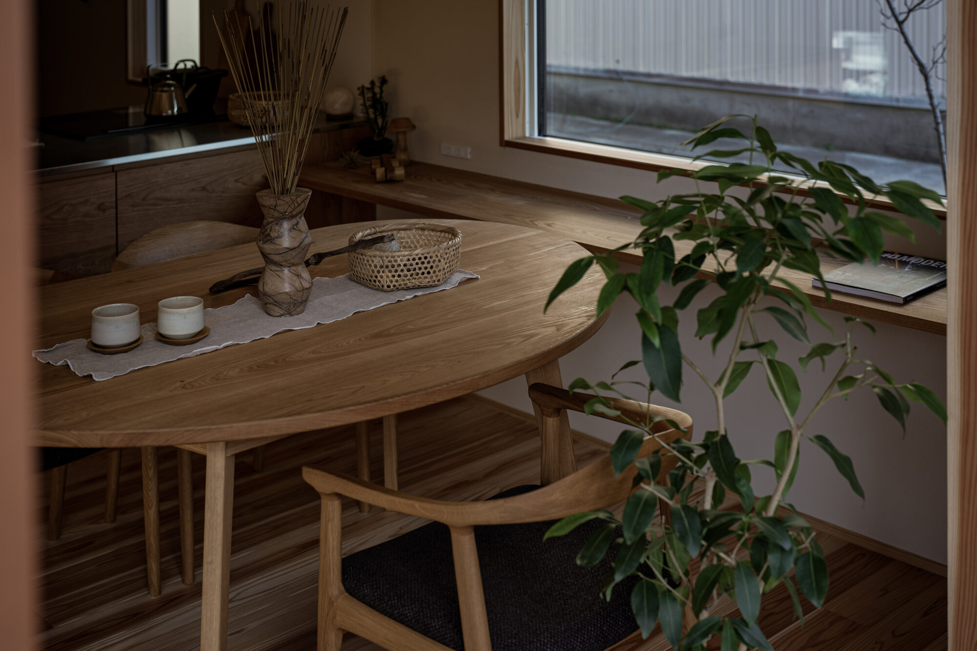 260217_096 - 滋賀で自然素材・木の家の注文住宅を建てる工務店|株式会社スムース Wooden dining table with woven basket centerpiece and candles, in a warm, cozy kitchen nook beside a window plant.