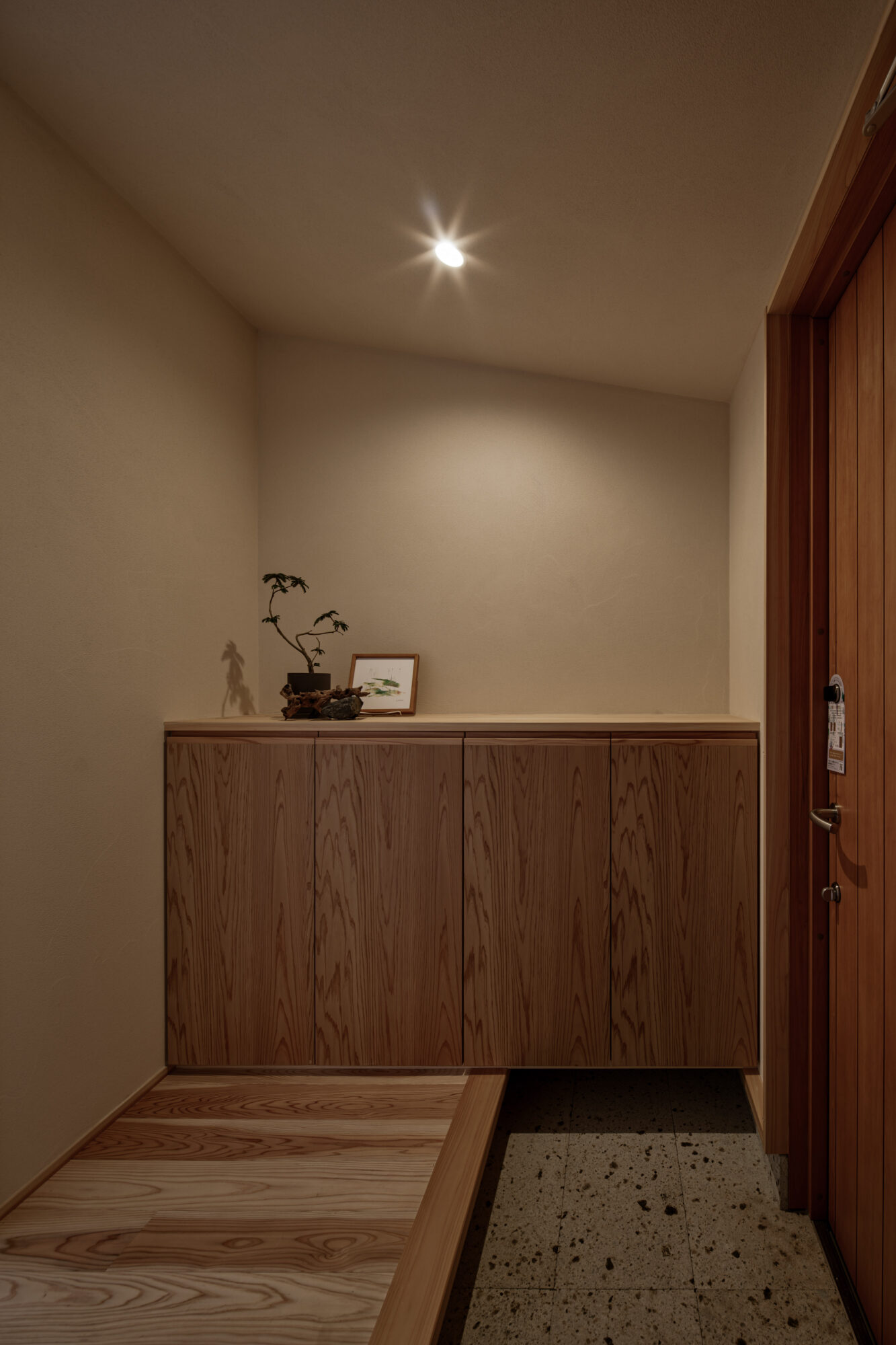260217_111 - 滋賀で自然素材・木の家の注文住宅を建てる工務店|株式会社スムース Beige-walled entry alcove with a long wooden cabinet, a small bonsai plant, and a framed picture on the countertop under a single ceiling light.