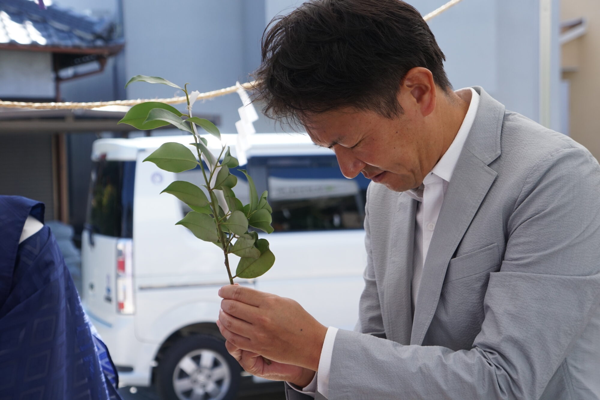 Man in a light gray suit bends to hand a leafy plant cutting to someone nearby outdoors, with a white van in the background.