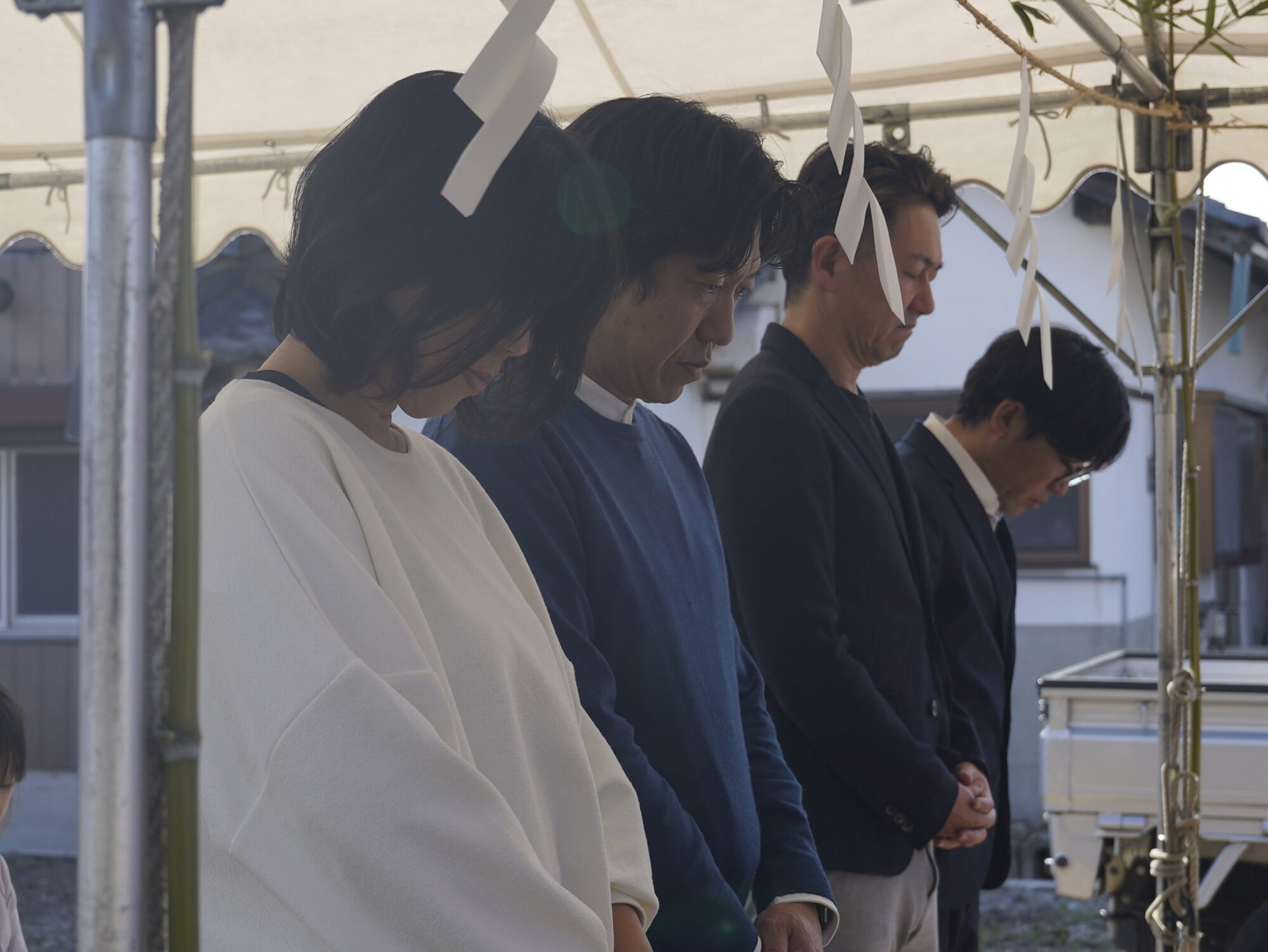 _DSC9887 1 - 滋賀で自然素材・木の家の注文住宅を建てる工務店|株式会社スムース People in formal clothing bow their heads in prayer under a canopy with white paper streamers hanging from a frame outside.
