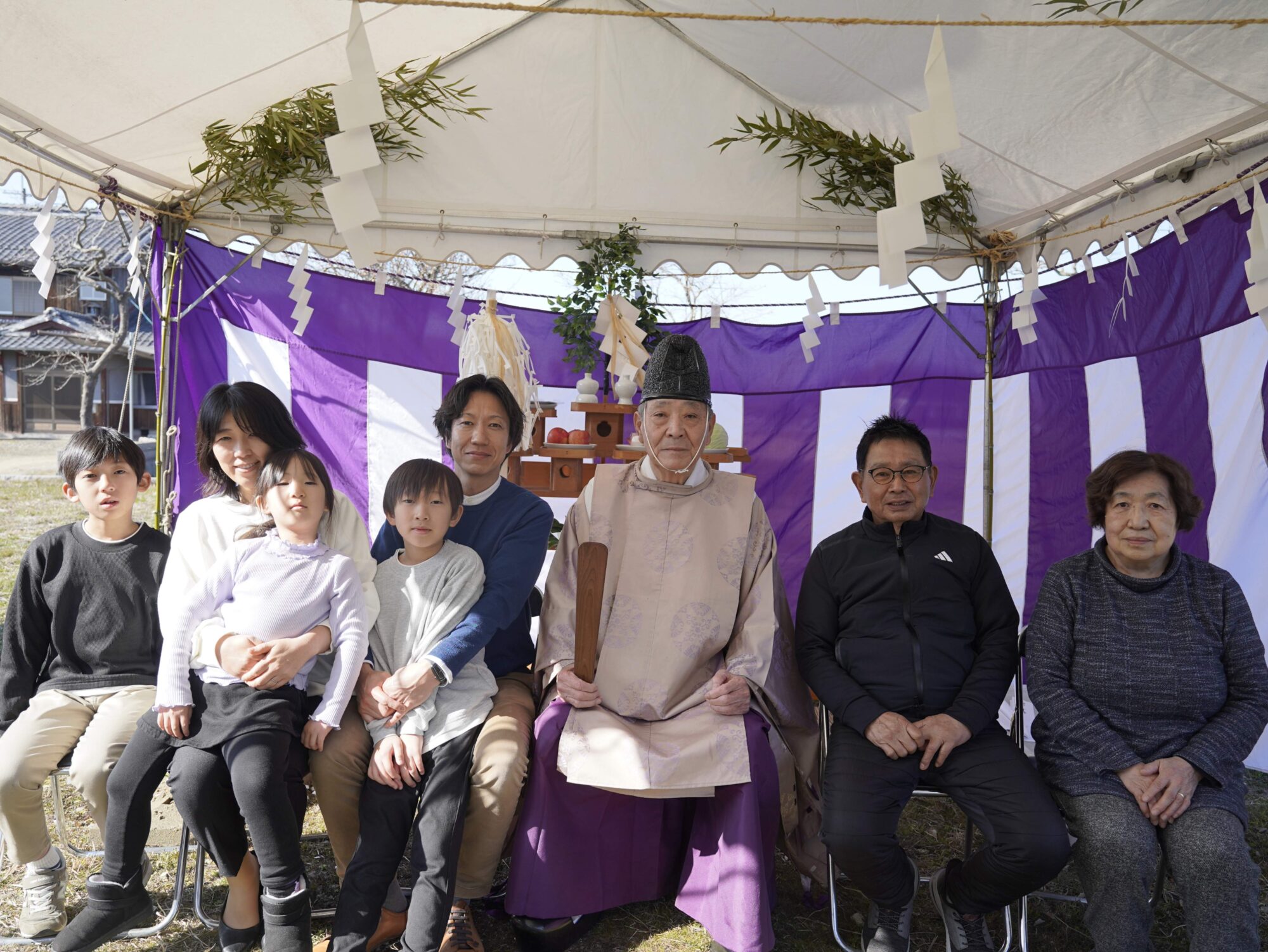 _DSC9950 1 - 滋賀で自然素材・木の家の注文住宅を建てる工務店|株式会社スムース Family and a priest pose for a group photo under a ceremonial tent with paper streamers and purple banners in the background.
